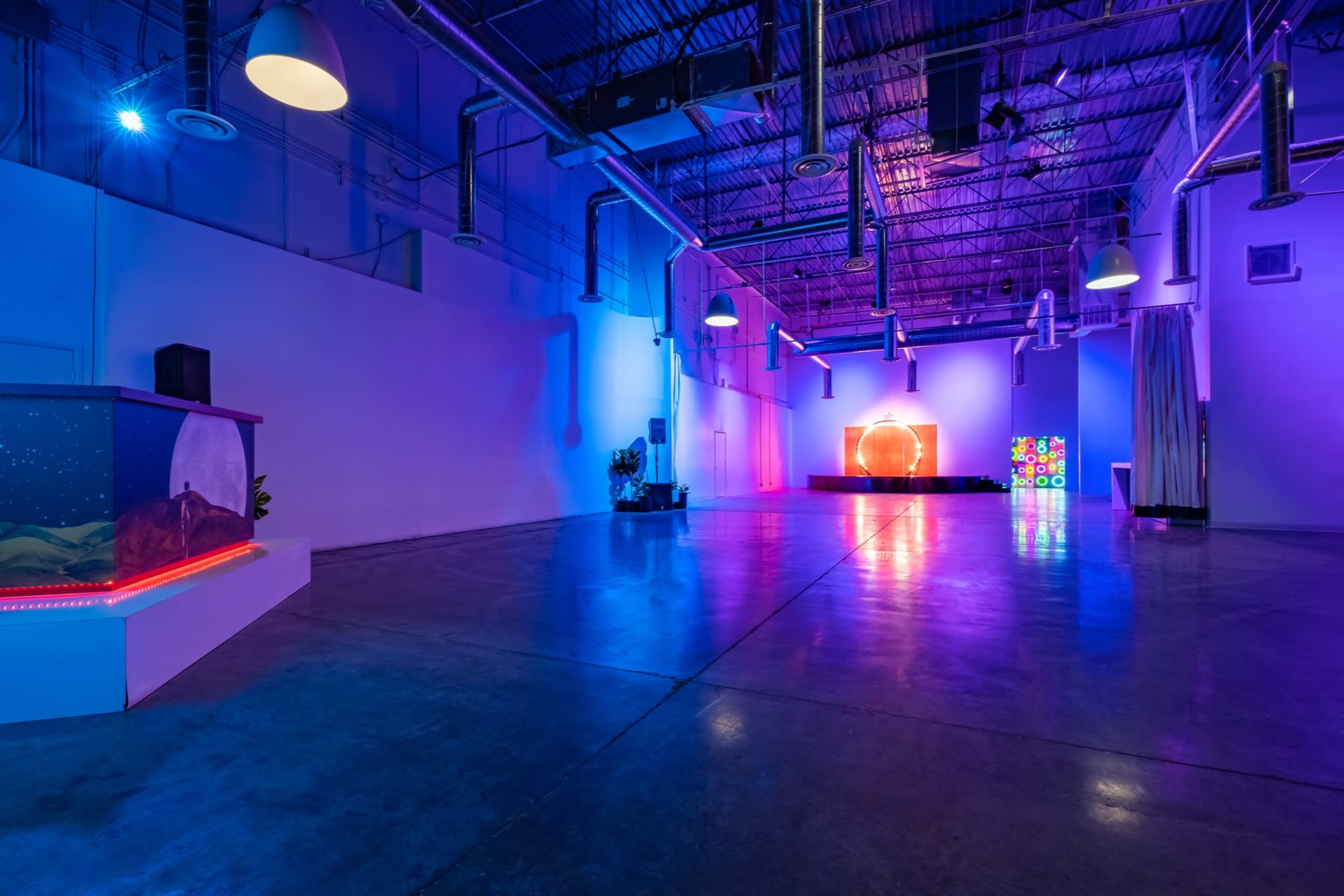A spacious art gallery features colorful lighting, polished concrete floors, and a stage with a large circular installation at one end.