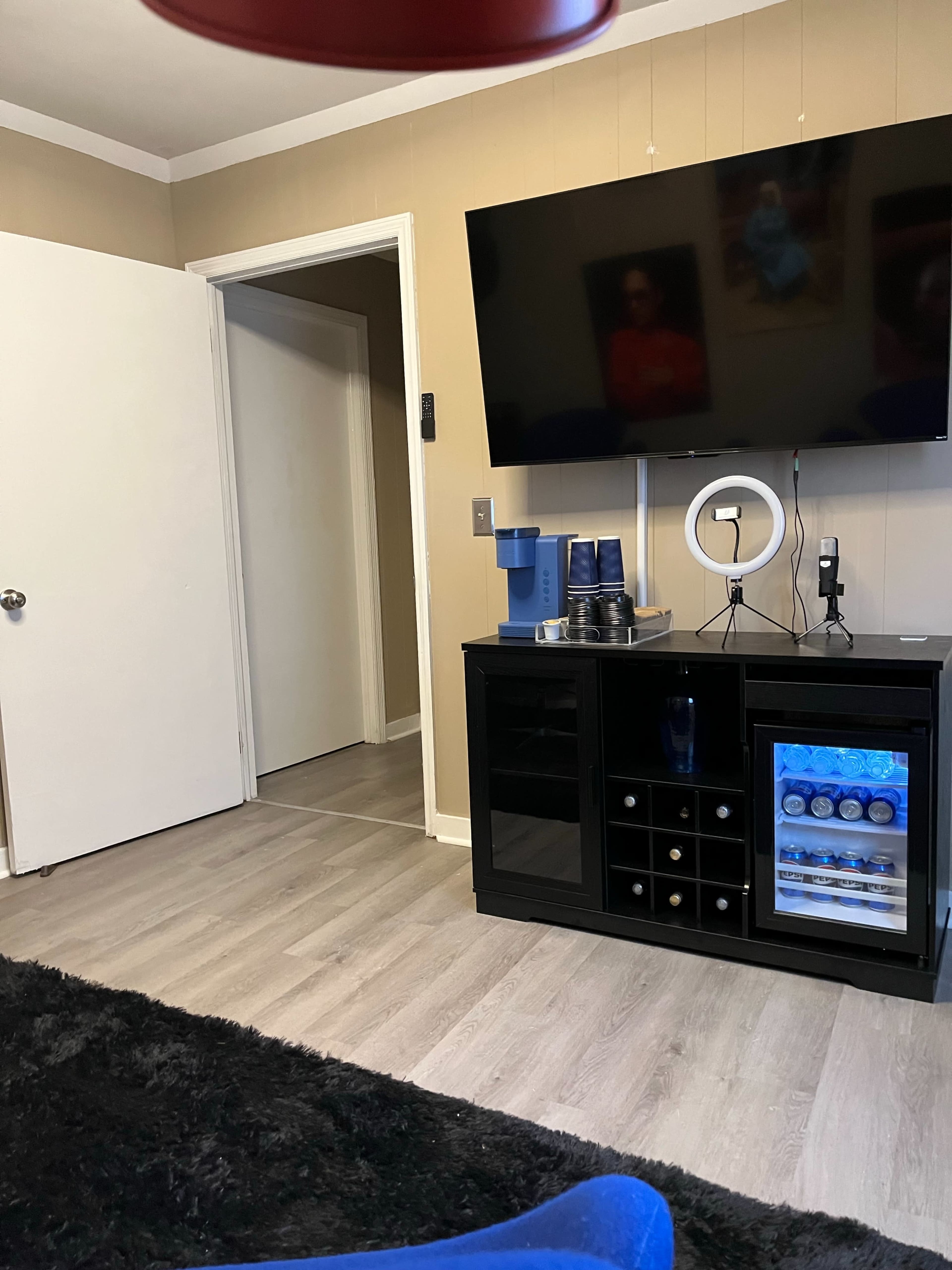 The image shows a living room with a black cabinet displaying a wine rack and a mini refrigerator, a TV mounted on the wall, and a doorway leading to another room.