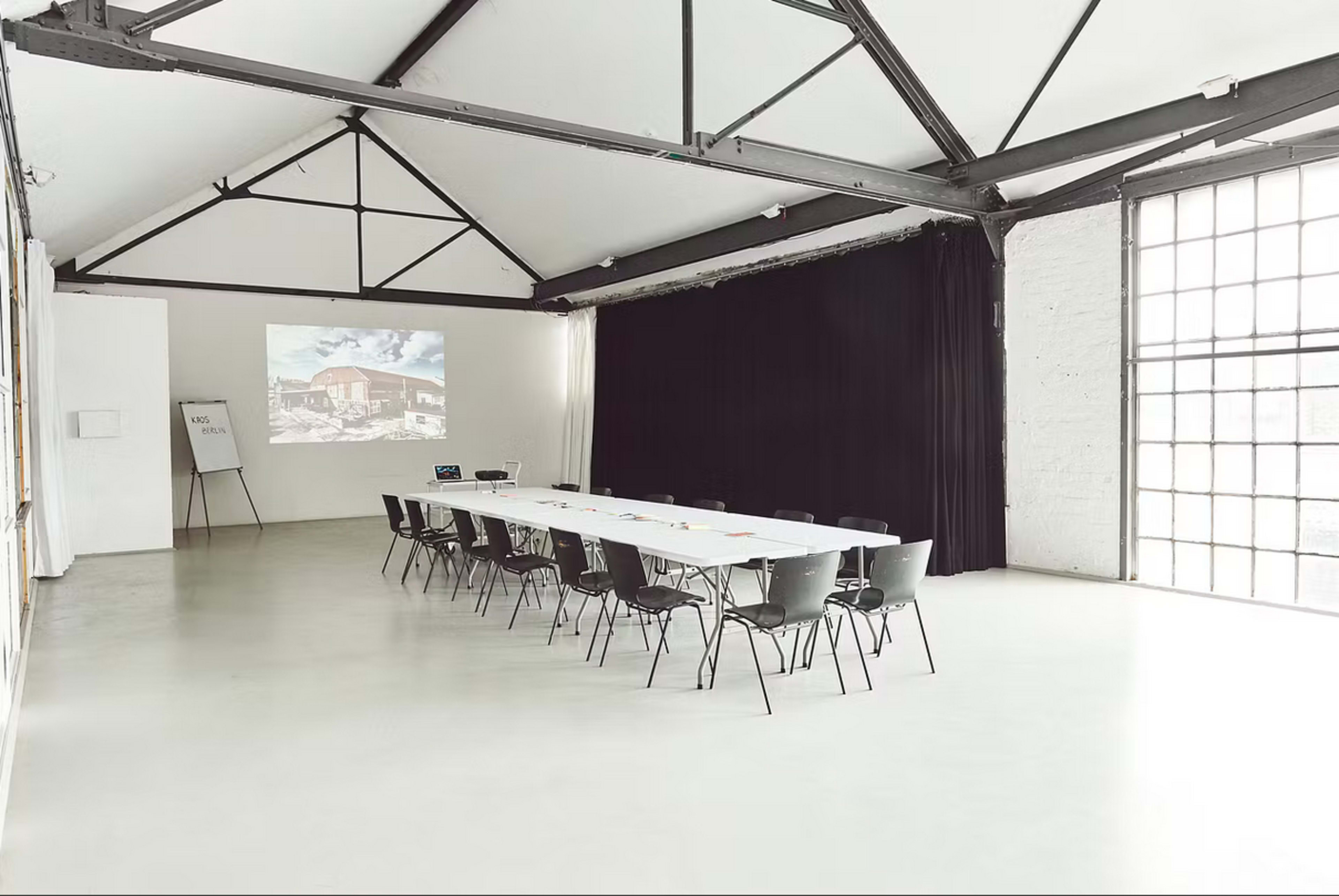 A spacious meeting room features a long table surrounded by chairs, with a projector display on one wall and large windows providing natural light.