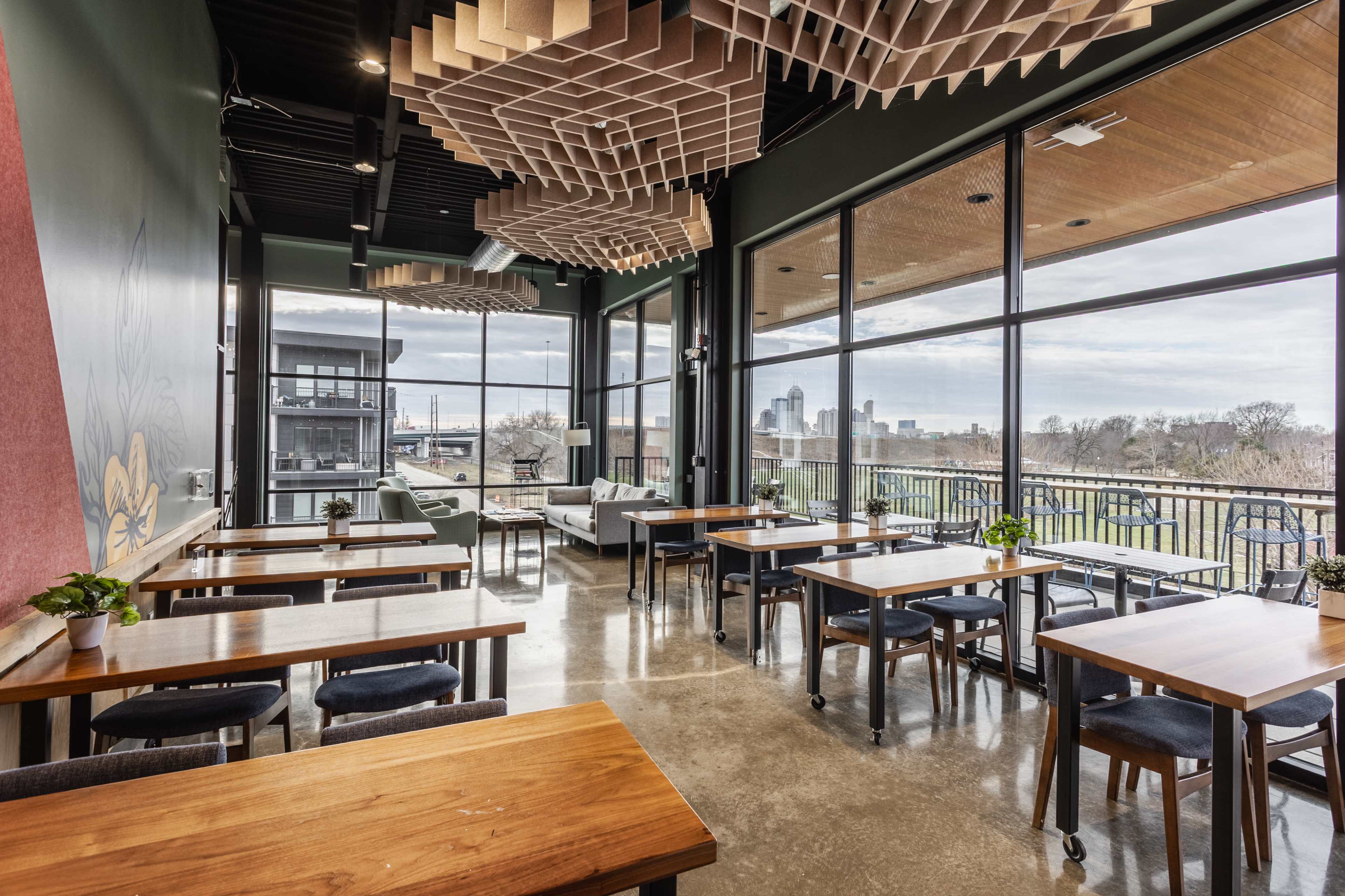 The image shows a modern restaurant interior with large windows offering a view of a city skyline, featuring wooden tables and contemporary decor.