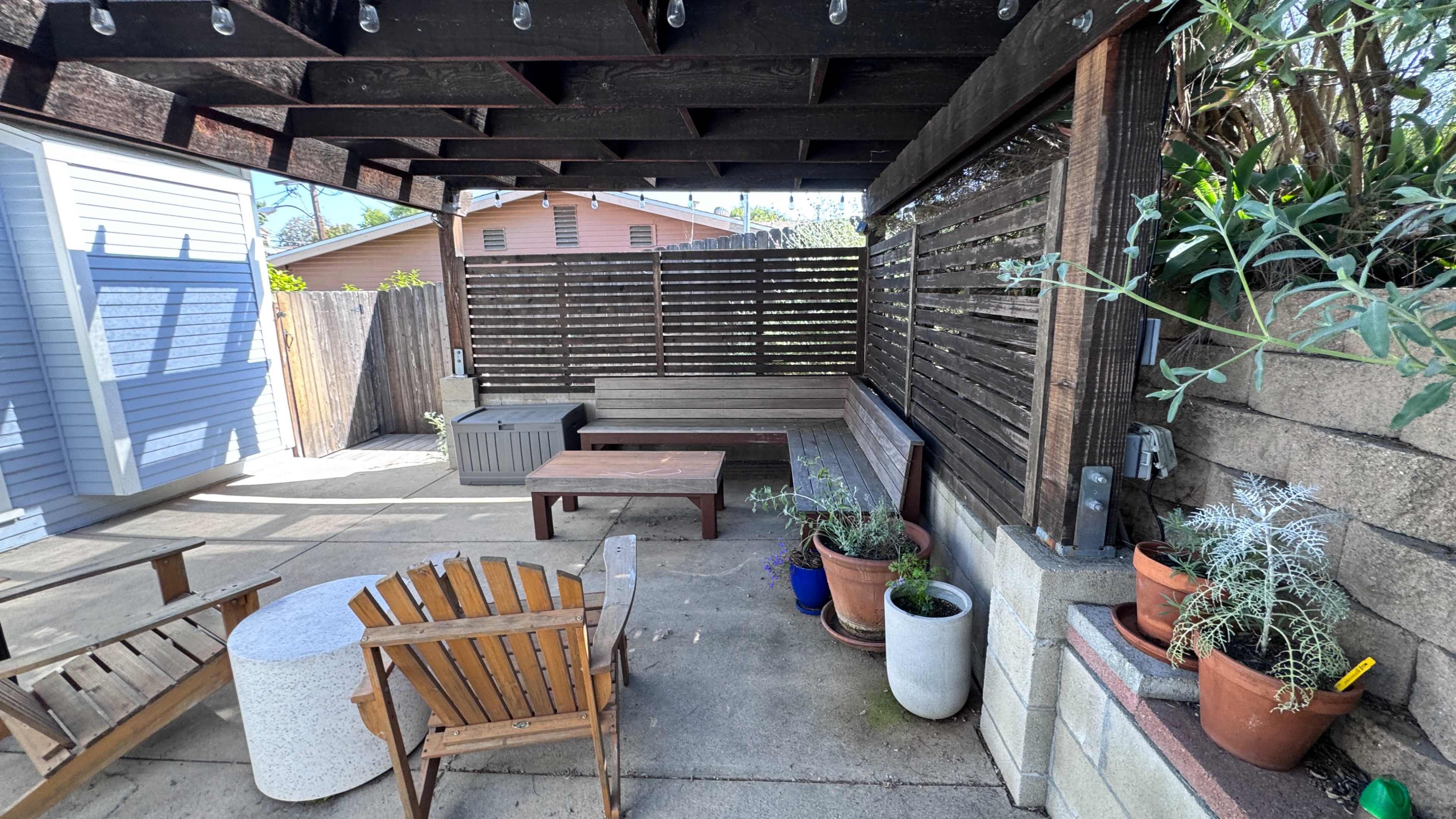 The image shows a cozy outdoor patio area with a wooden pergola, seating and planters, and a small table.