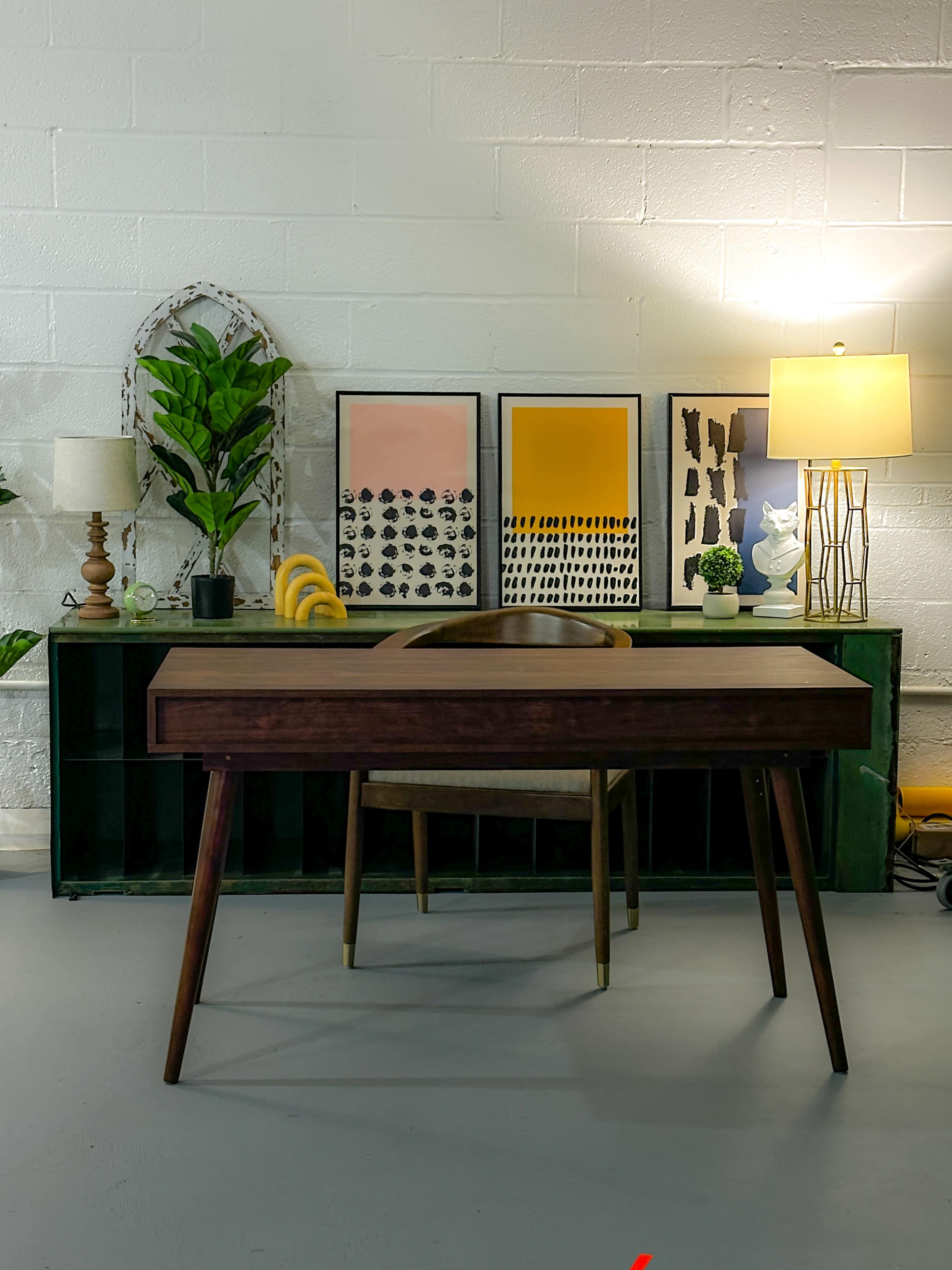 A modern wooden desk is positioned in front of a green console table adorned with potted plants and framed artwork, while a lamp casts light in the background.
