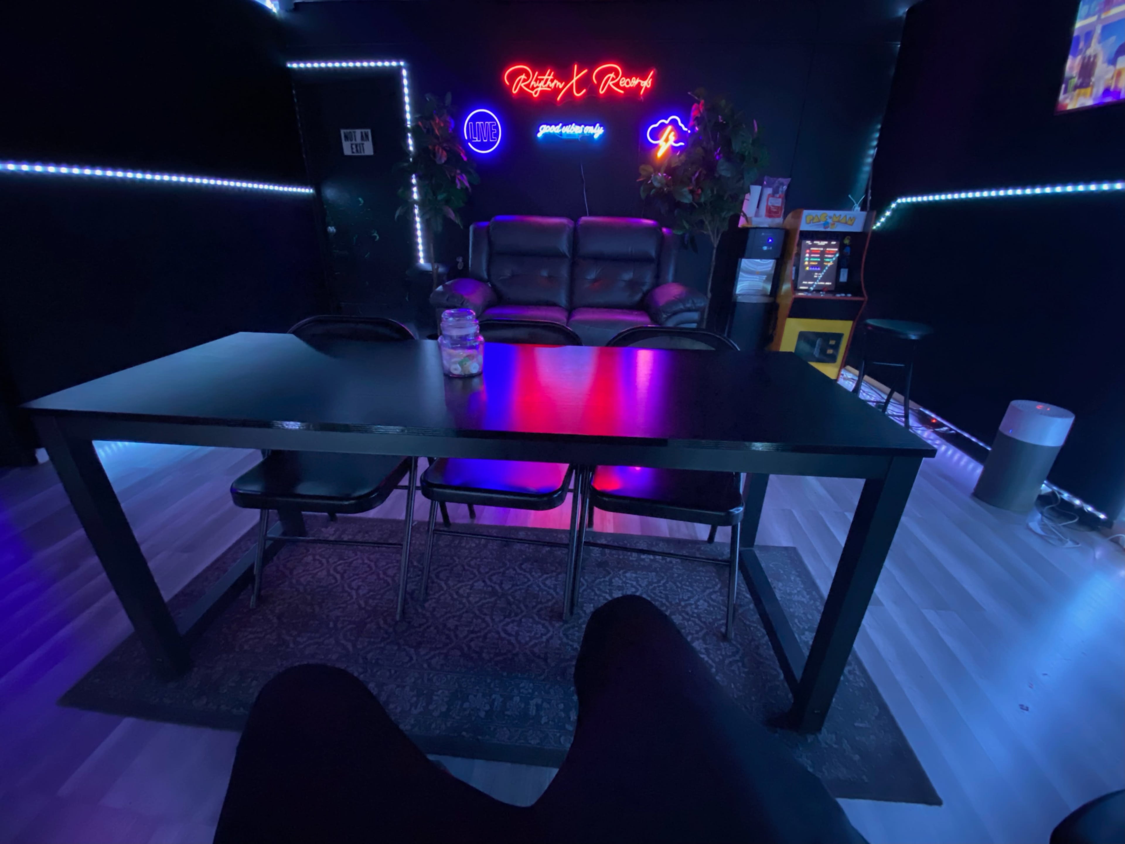 A dimly lit room with a black table and four chairs, a couch in the background, and neon lights illuminating the walls.