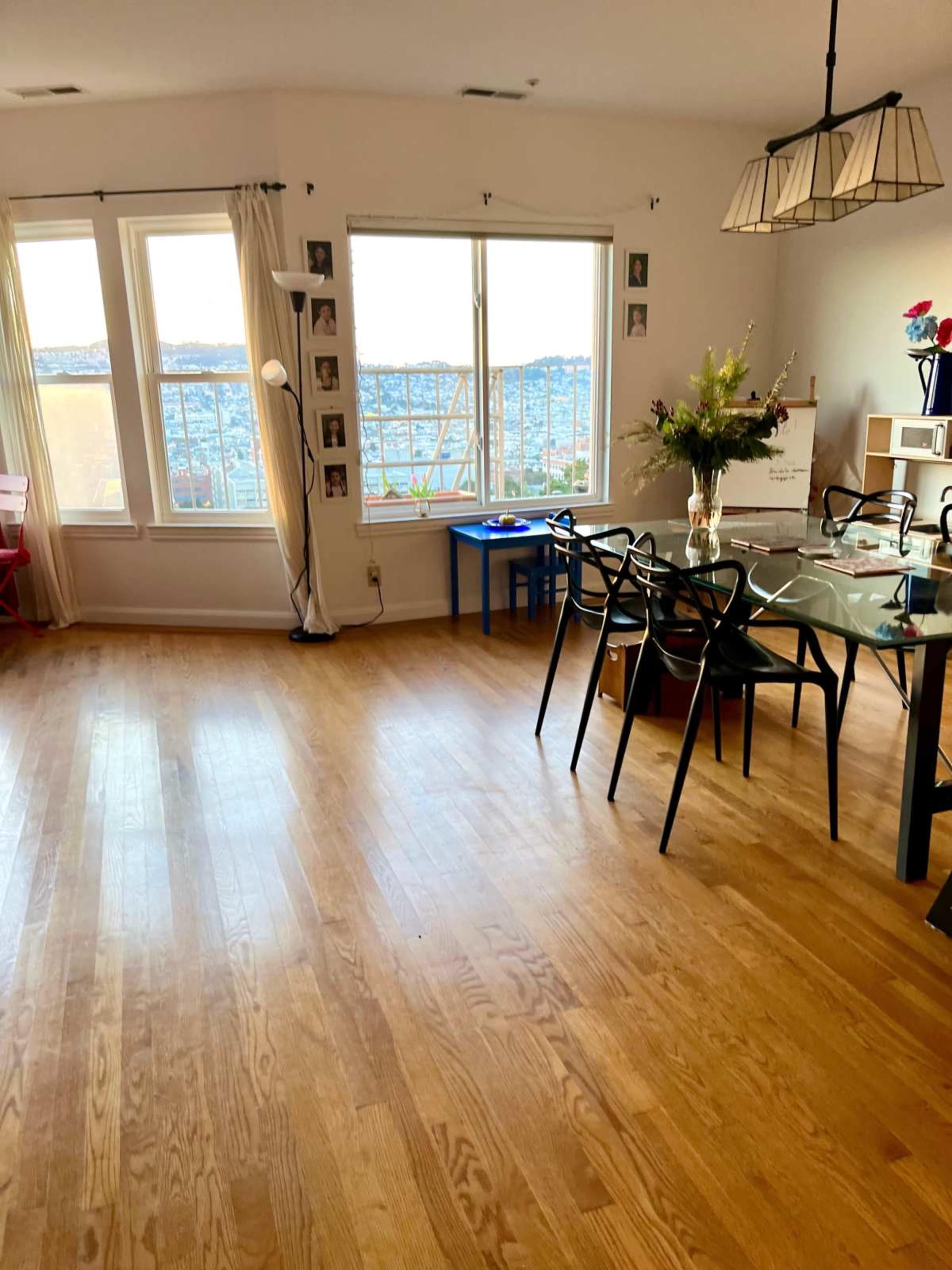 The image shows a bright, open living space with wooden flooring, large windows offering a view, a dining table with black chairs, and a small table in the corner.