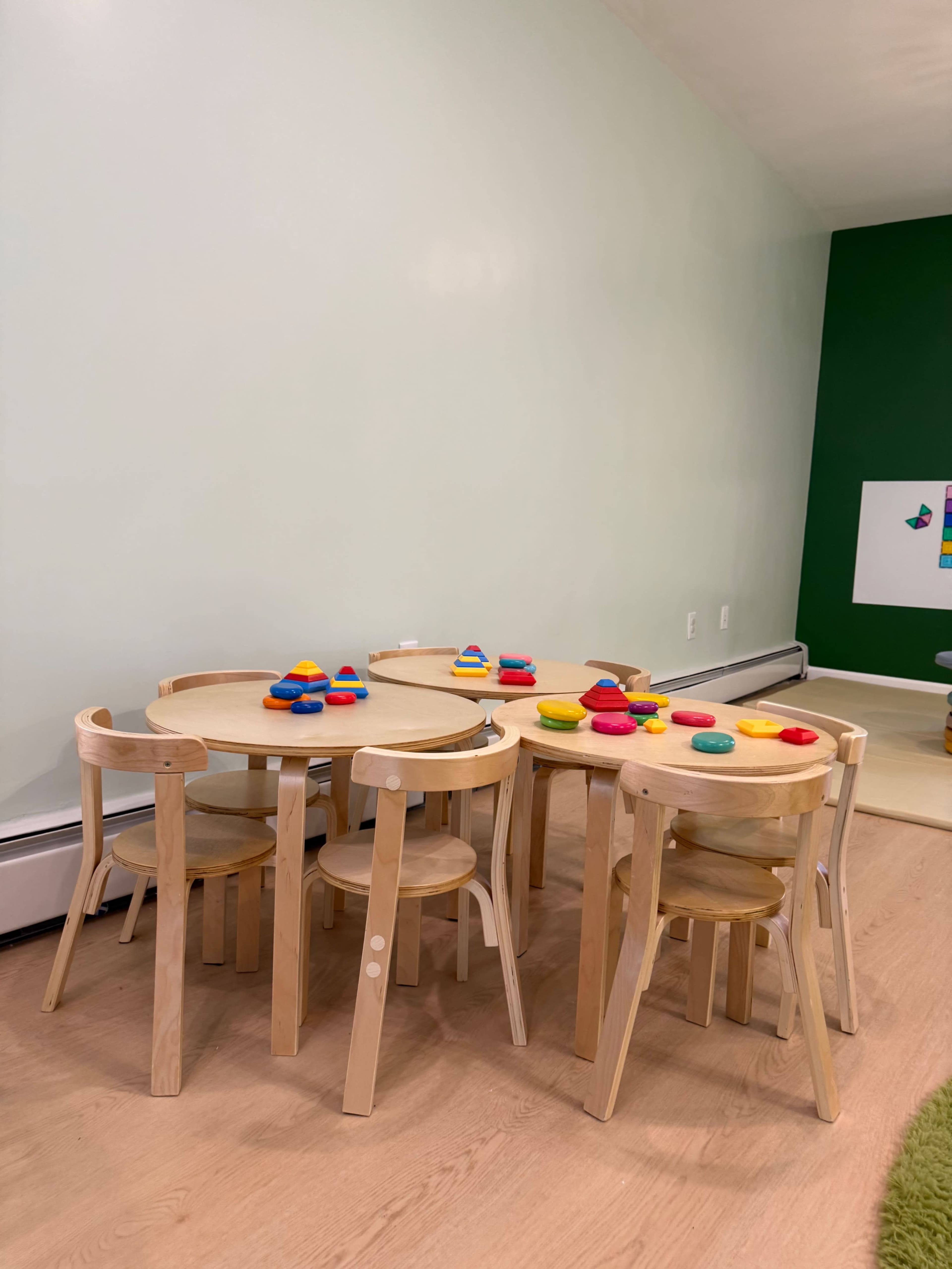 The image shows a play area with two wooden tables surrounded by matching chairs, featuring colorful toys on the surfaces.