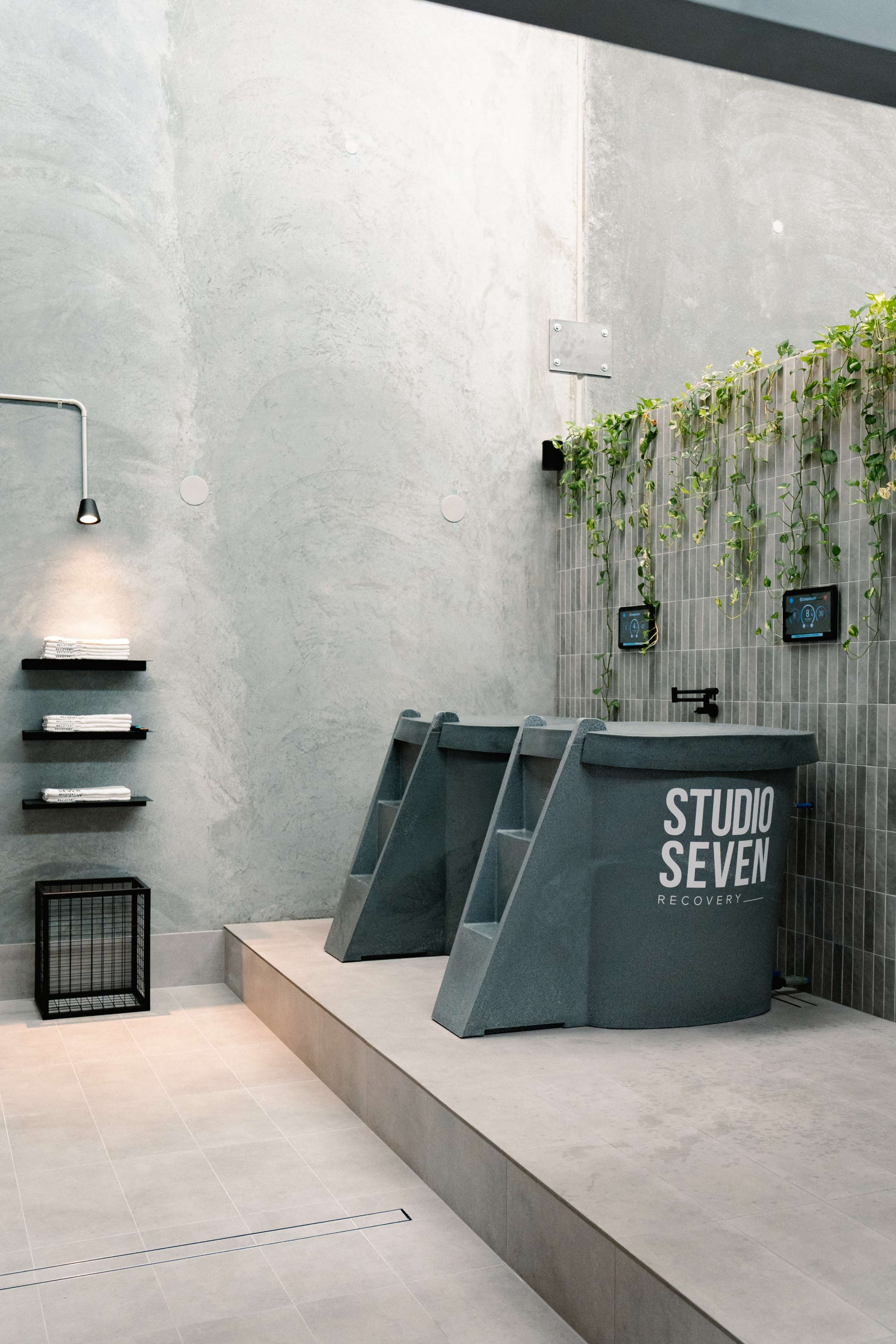 The image shows a modern recovery room featuring a pair of gray tubs, a green wall with plants, and minimalist shelving against a textured gray wall.