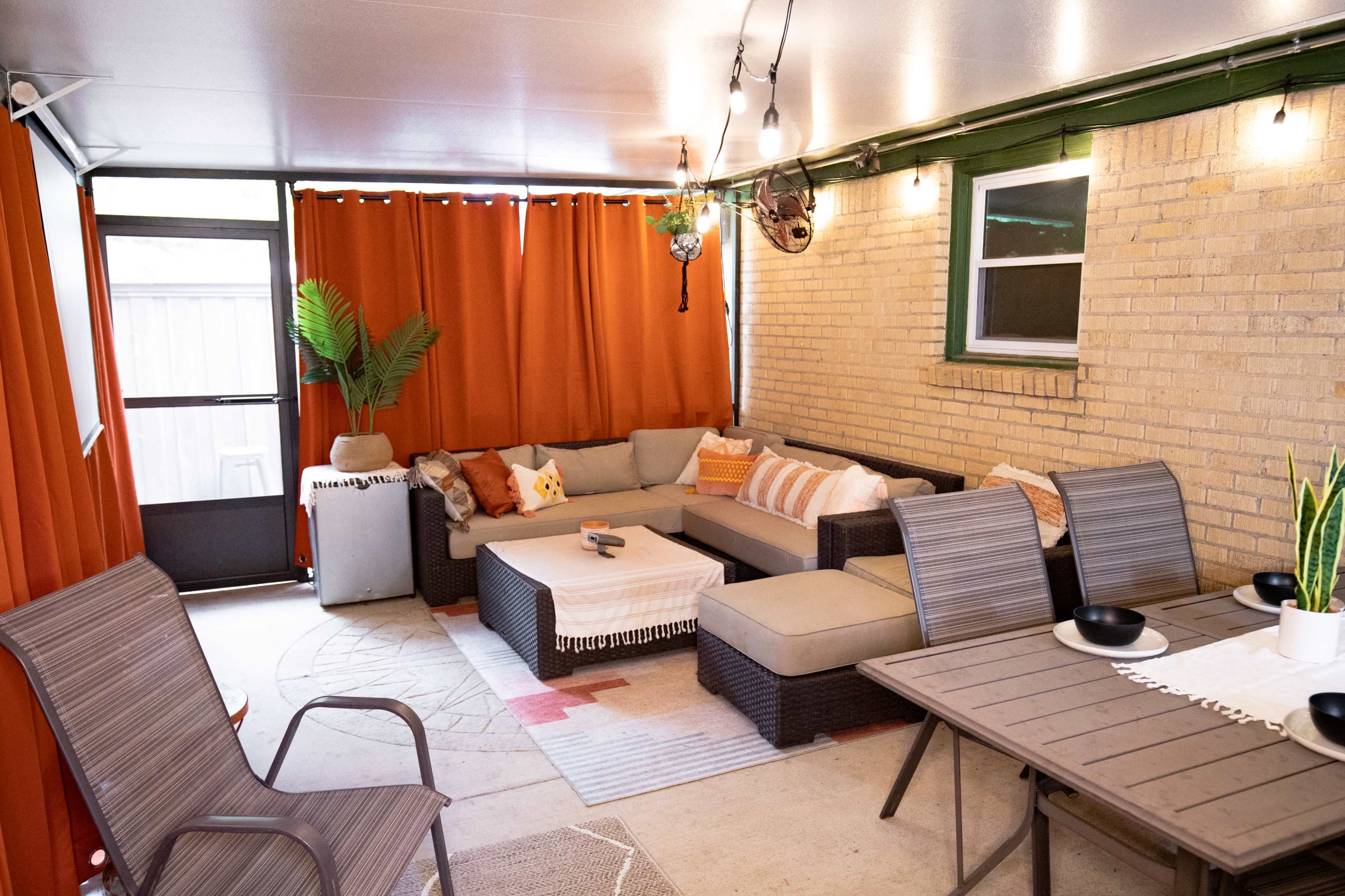 A cozy, covered patio area featuring a sectional sofa, dining table, and decorative plants, with orange curtains and string lights accentuating the space.