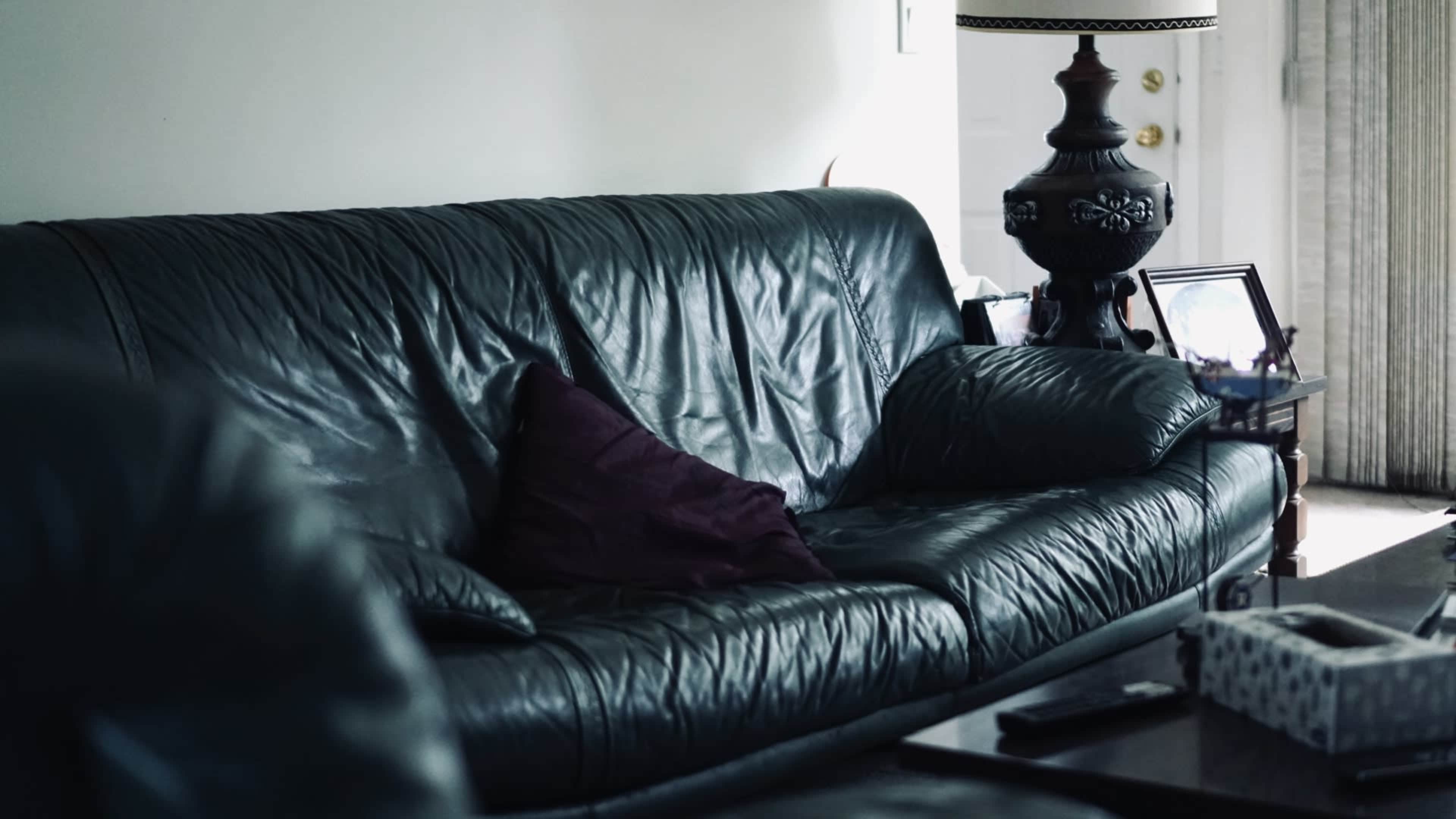 The image shows a black leather sofa with a purple throw pillow, positioned near a lamp and a table.