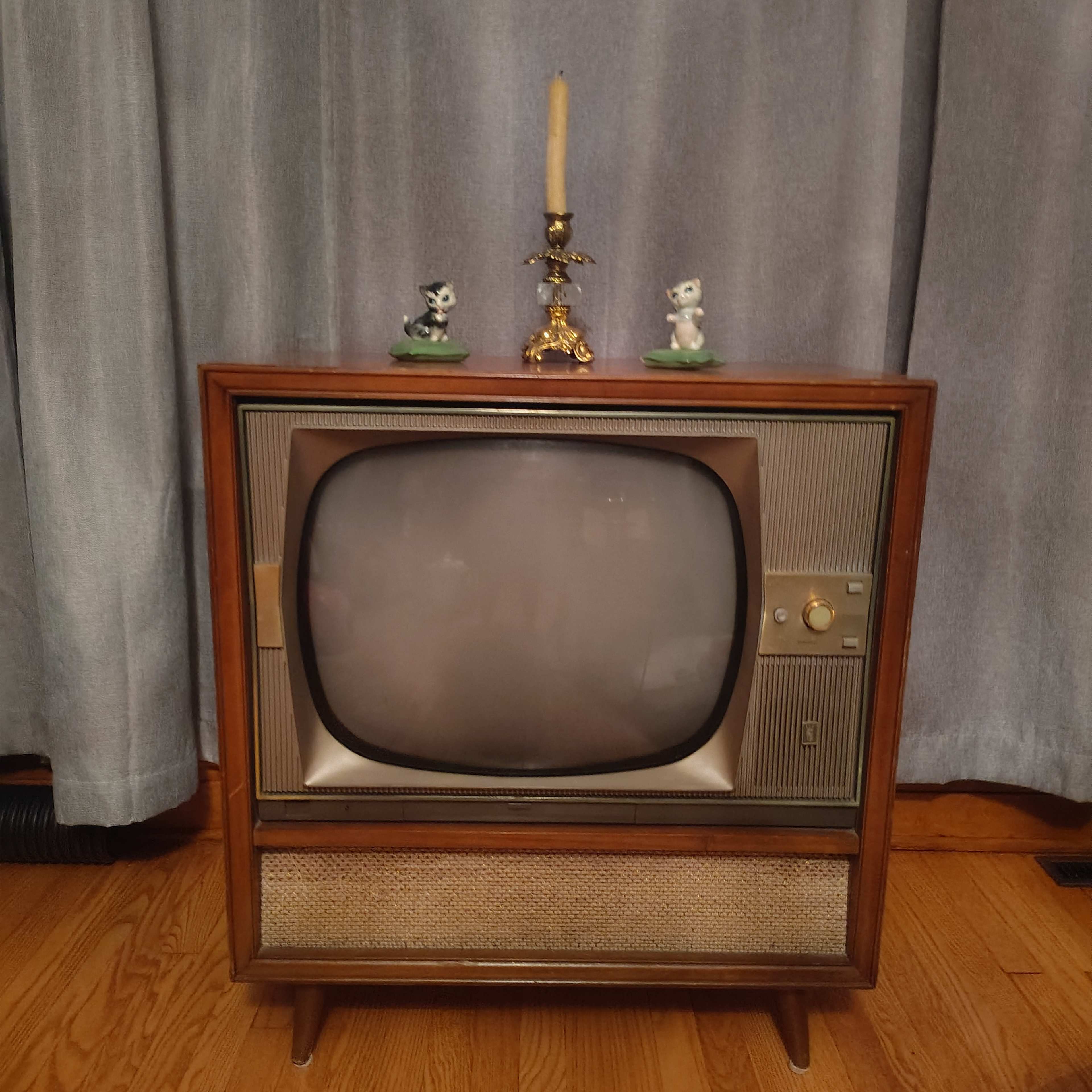 The image shows a vintage television set with a rounded screen, placed in front of a light blue curtain, and adorned with a candlestick and two small figurines on top.