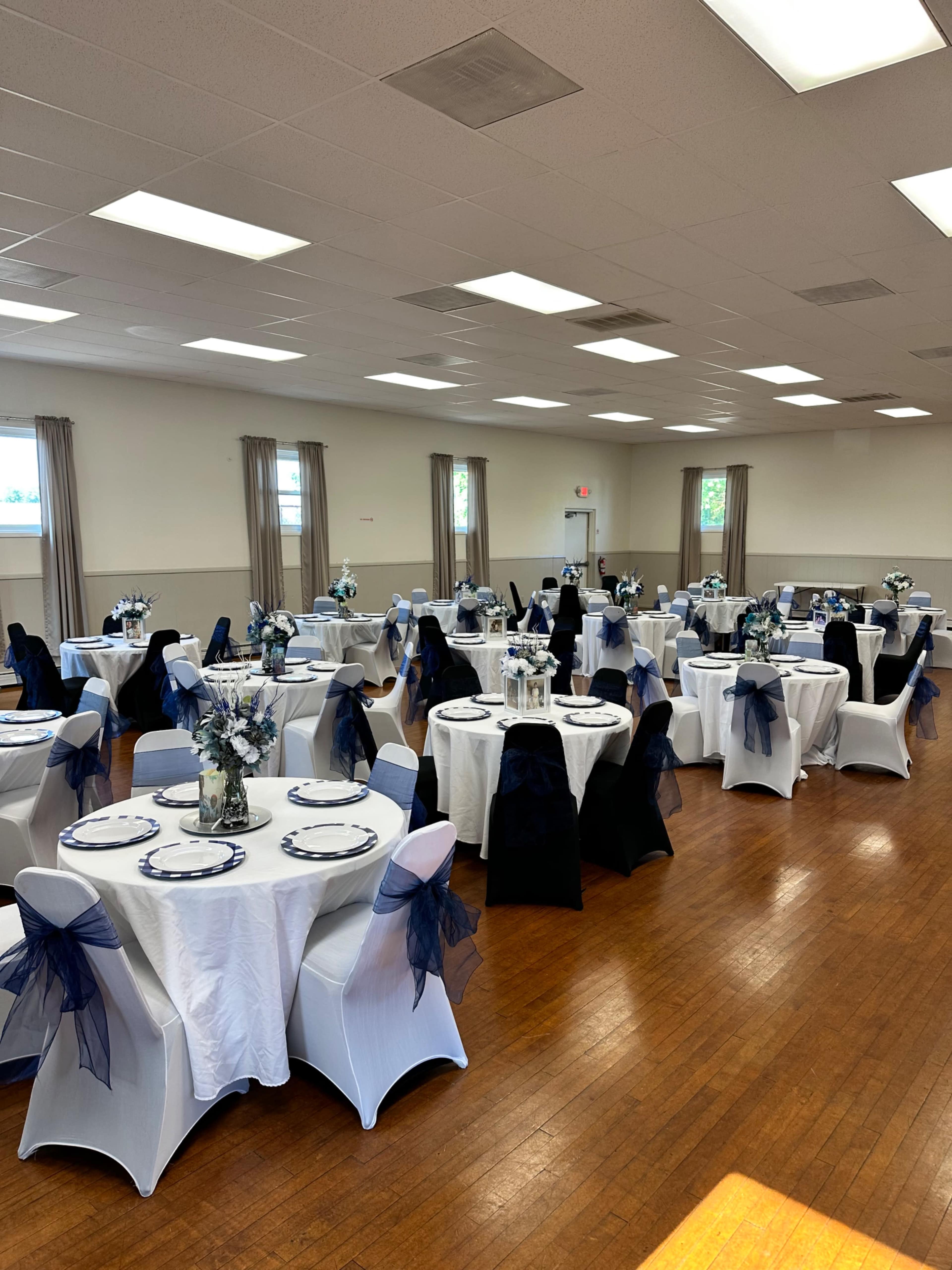 The image shows a banquet hall with round tables dressed in white tablecloths and dark blue decorations, arranged neatly across the wooden floor.