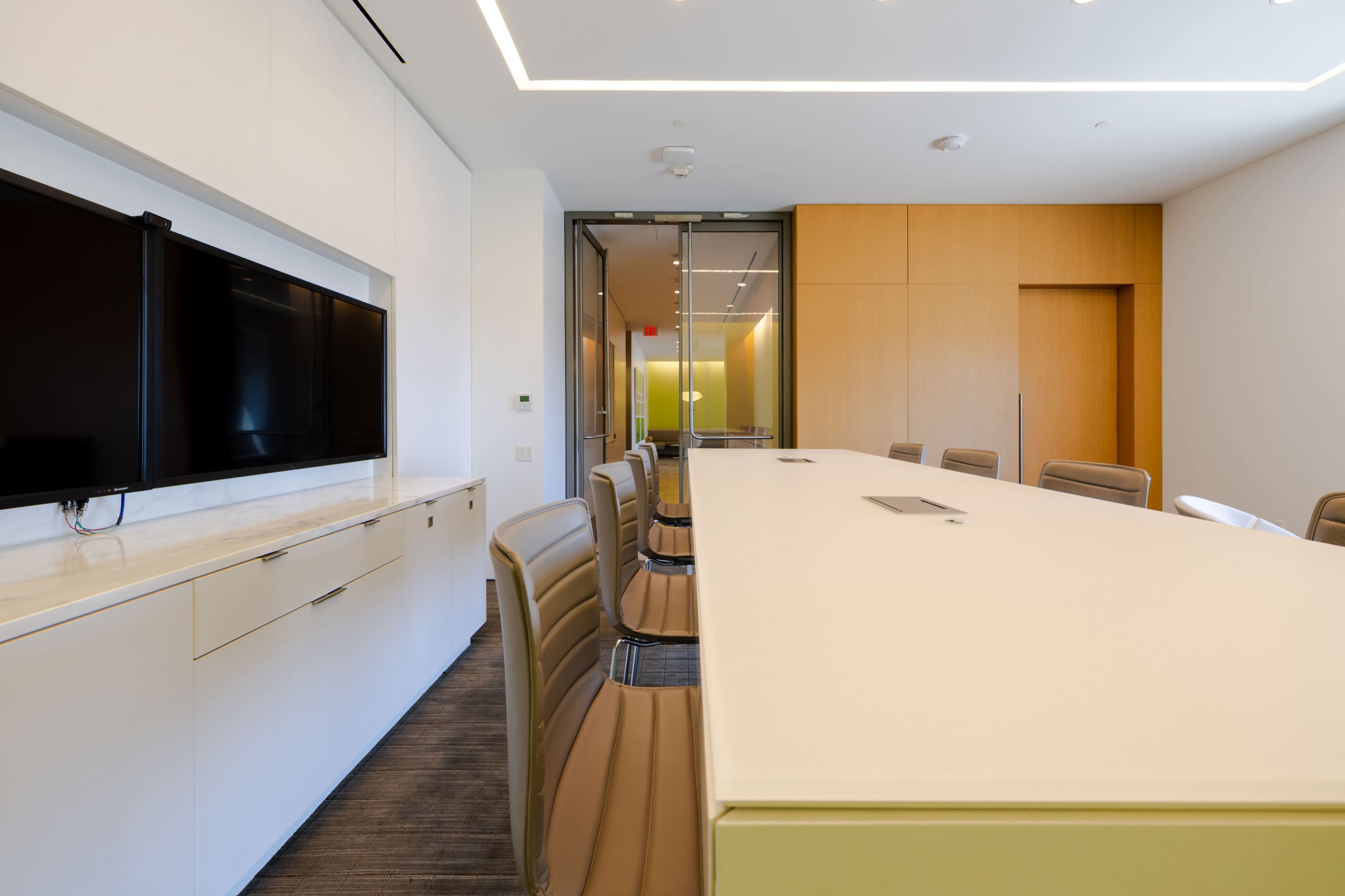 The image shows a modern conference room with a long table, chairs, a television mounted on the wall, and glass doors leading to another space.