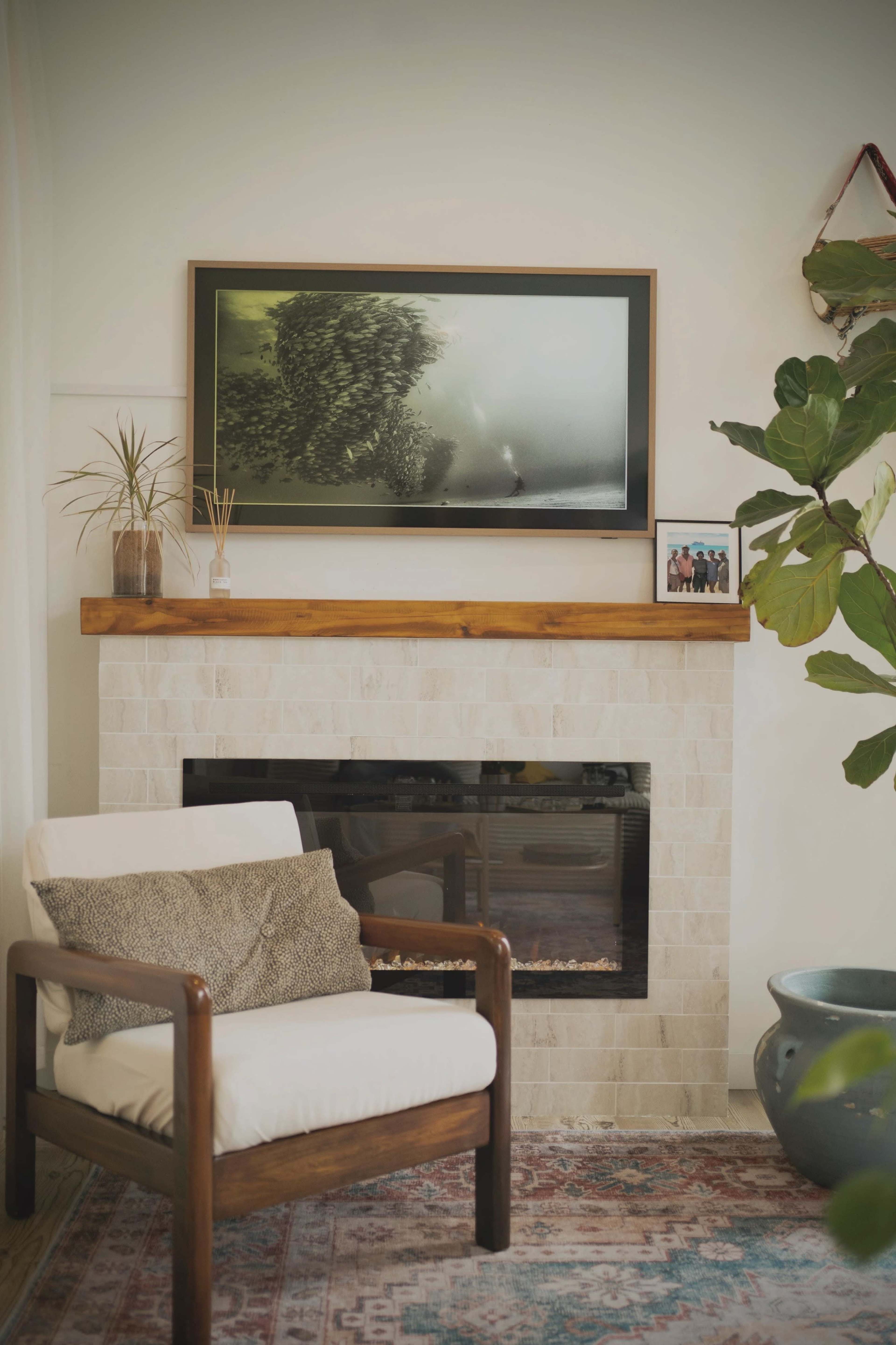 A cozy living room features a wooden-framed chair, a fireplace with a white stone surround, a large framed photograph above the mantel, and a decorative plant nearby.