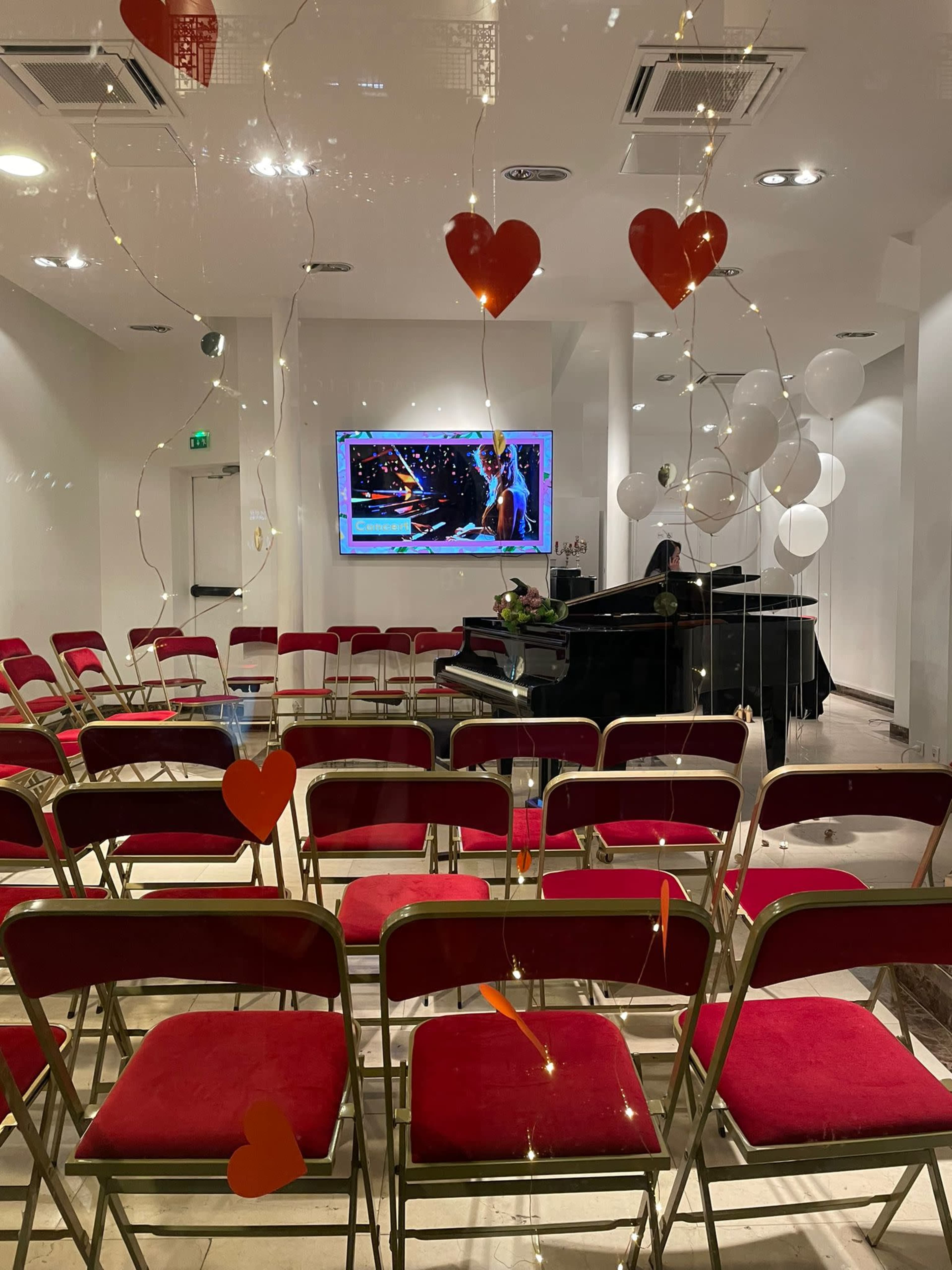 The image shows a staged indoor event space with rows of red chairs facing a piano and a screen displaying a performance, decorated with heart-shaped balloons and string lights.