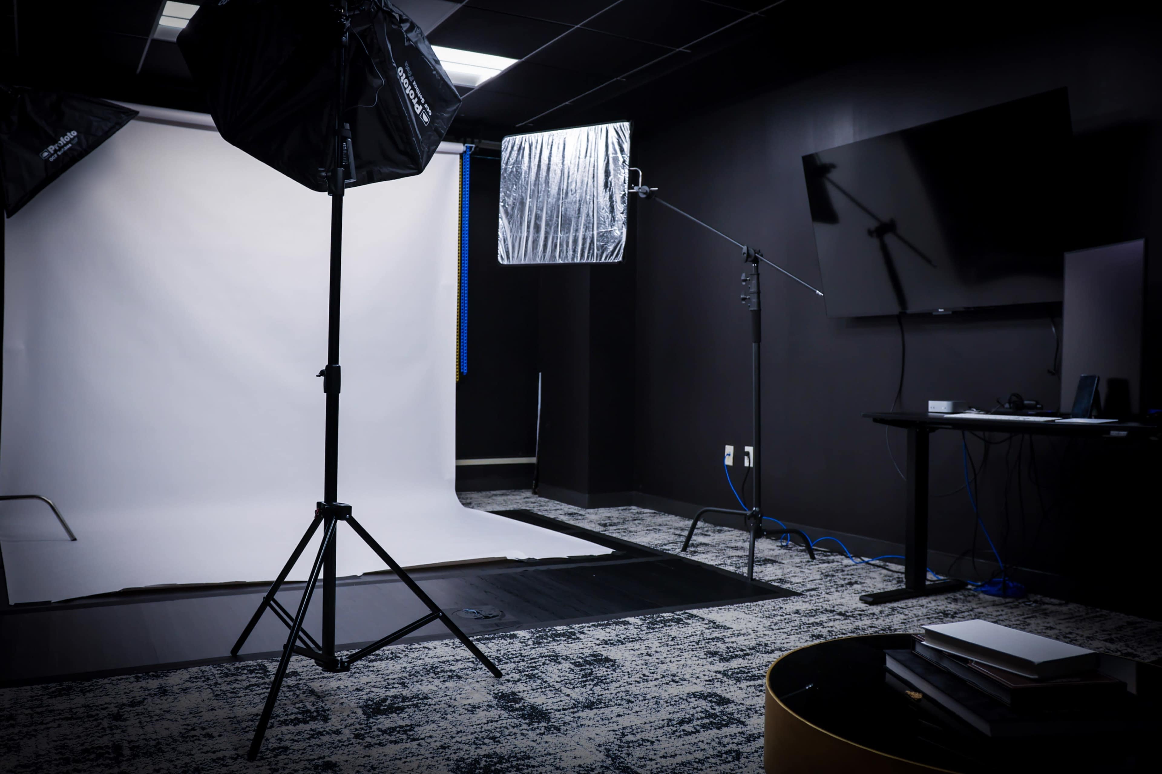 The image shows a photography studio setup with a white backdrop, lighting equipment, and a dark room containing a television and a desk with electronic devices.
