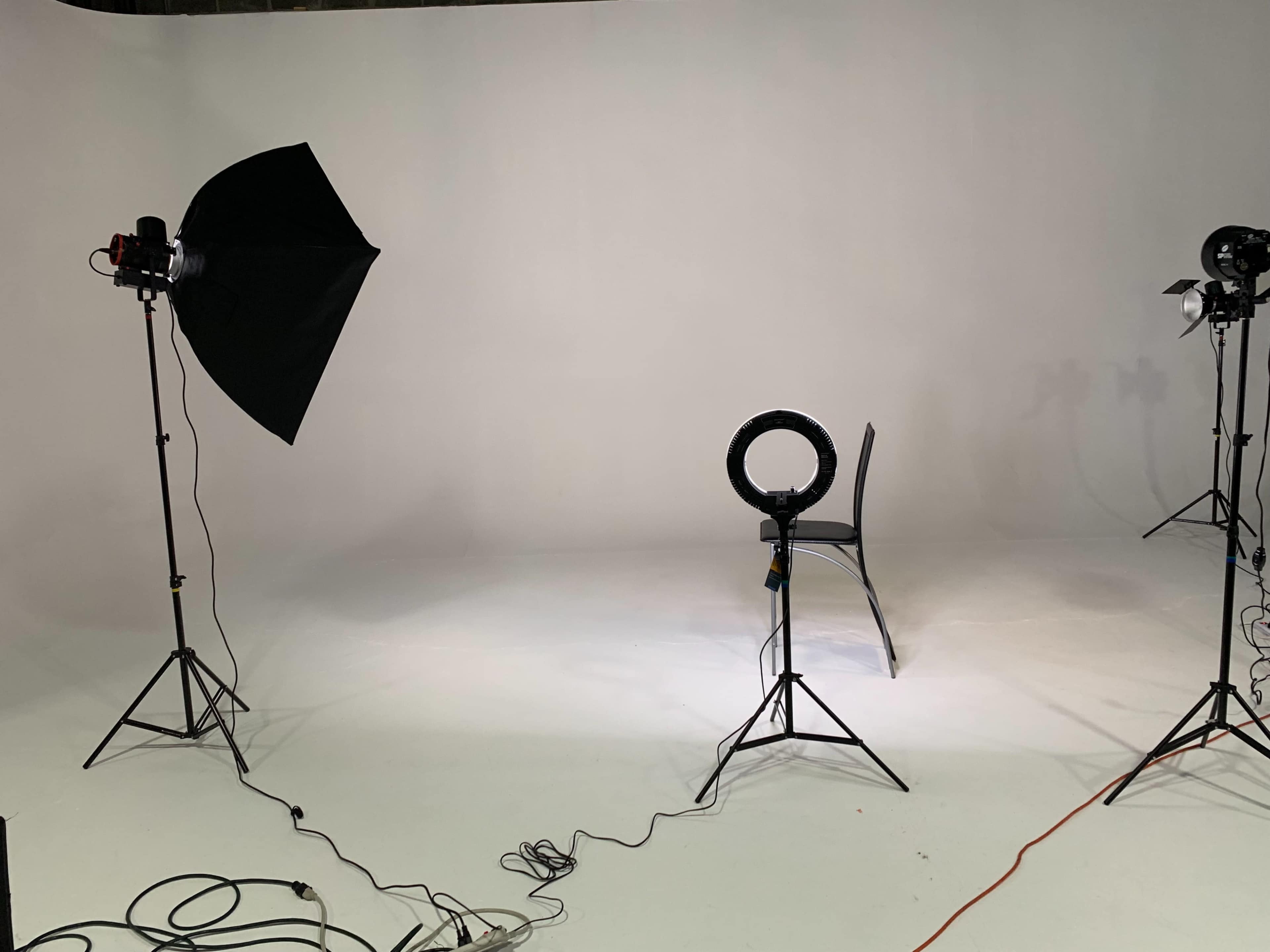 A photography studio is set up with a chair in the center, surrounded by multiple lighting equipment on stands, against a plain white background.