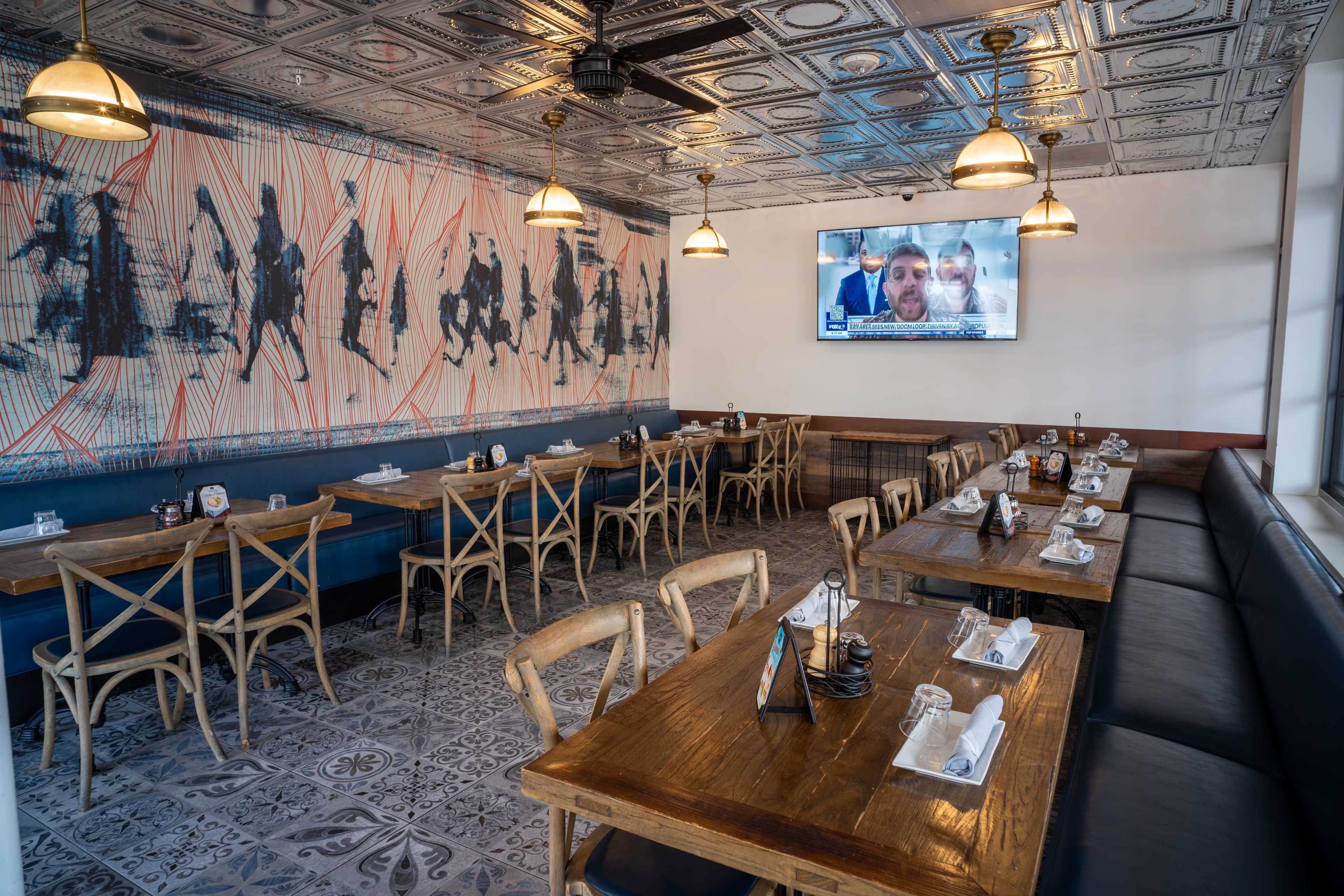 The image shows a dining area in a restaurant with wooden tables, chairs, and a large wall mural, along with a television displaying news.