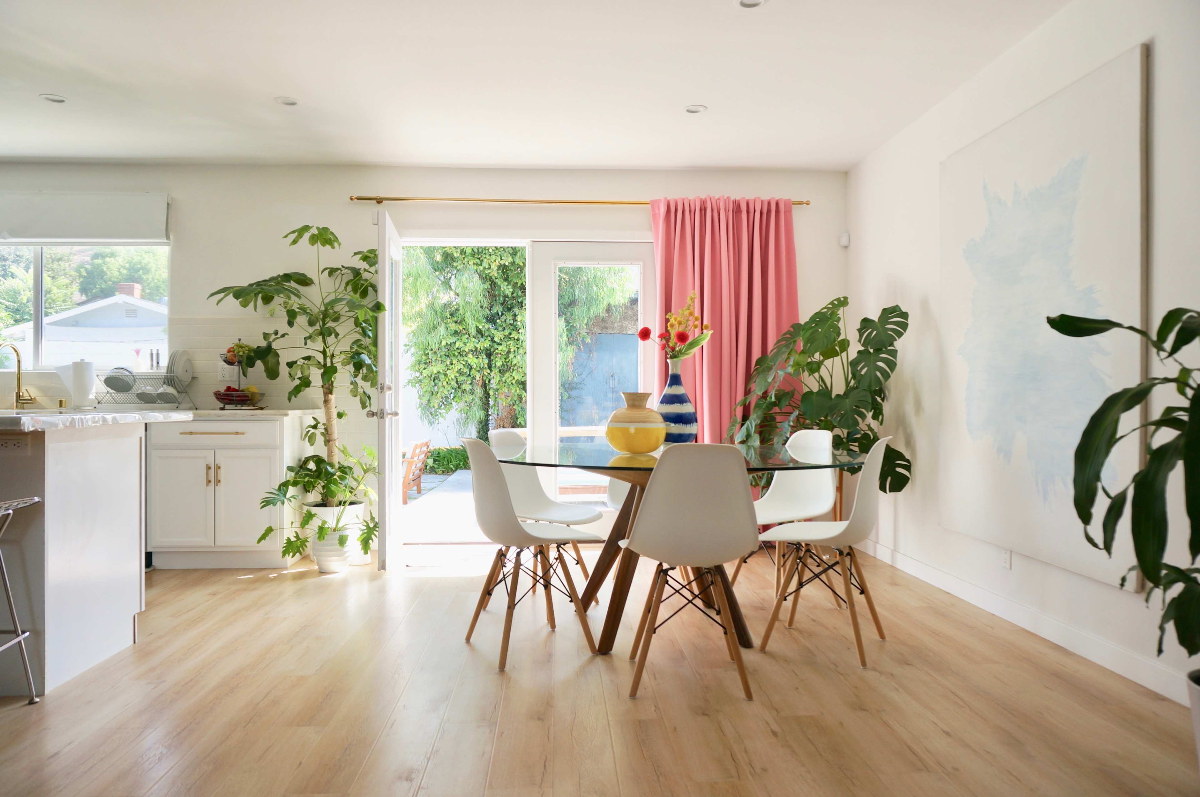 A bright dining area features a glass table surrounded by white chairs, potted plants, and a large artwork on the wall.