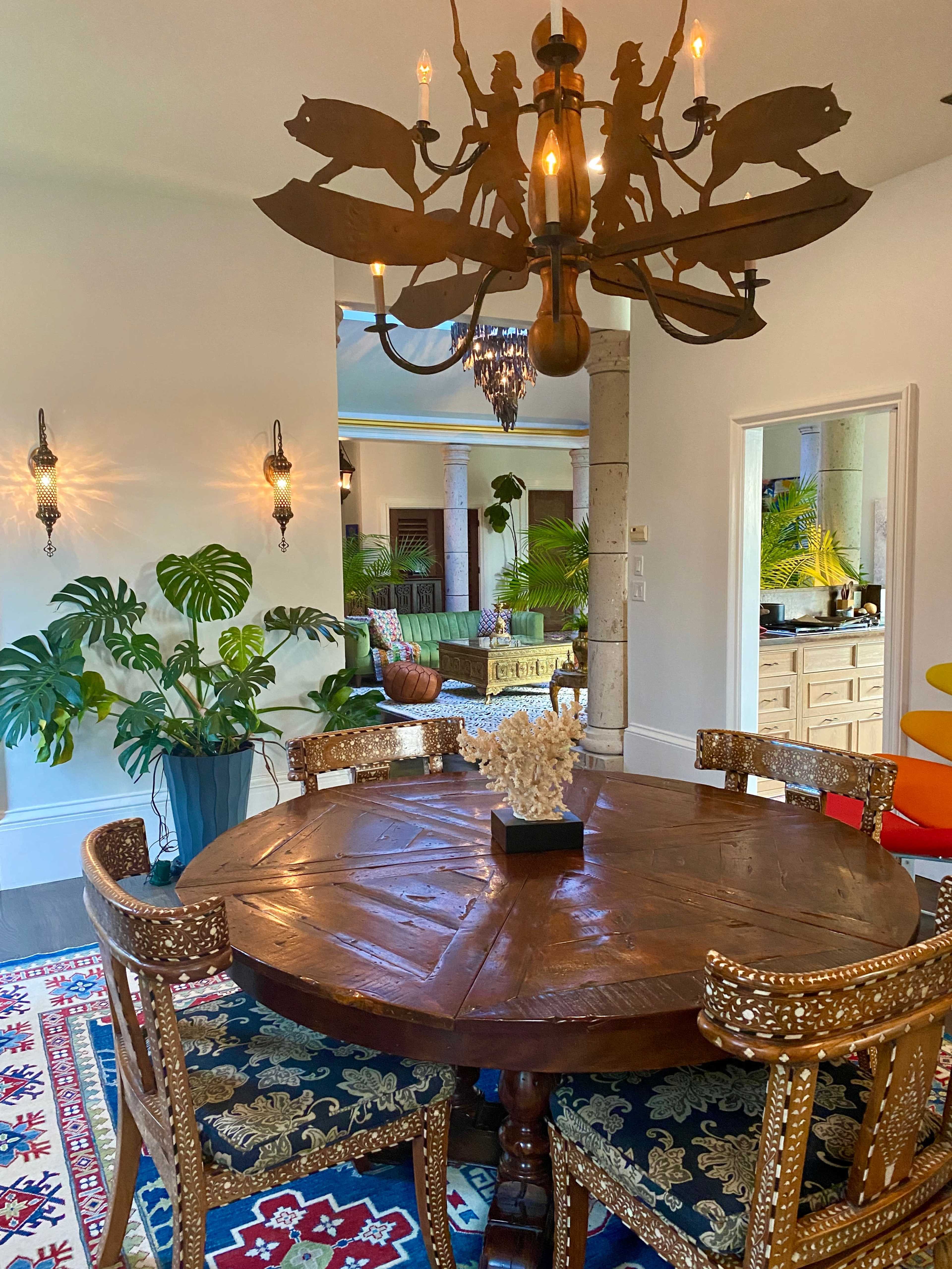The image shows a round wooden dining table surrounded by intricately designed chairs, with a chandelier featuring horse figures hanging above, and a glimpse of a brightly colored living area through an archway.