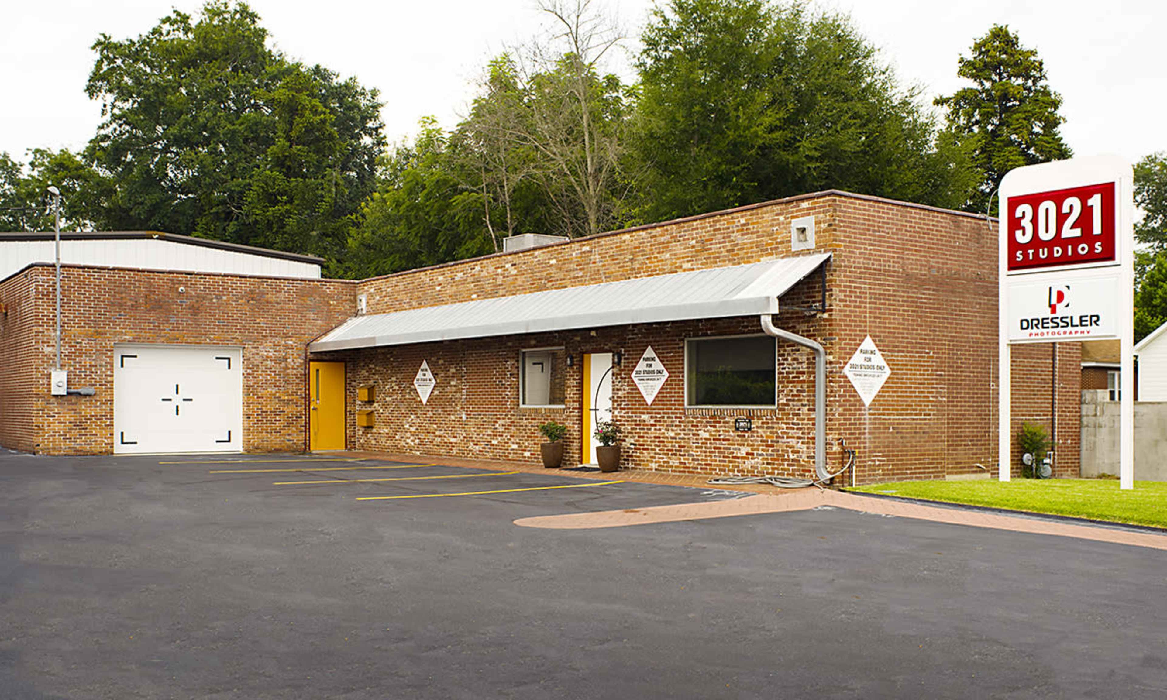 A brick building labeled "3021 Studios" with a sign and a parking area, surrounded by trees.