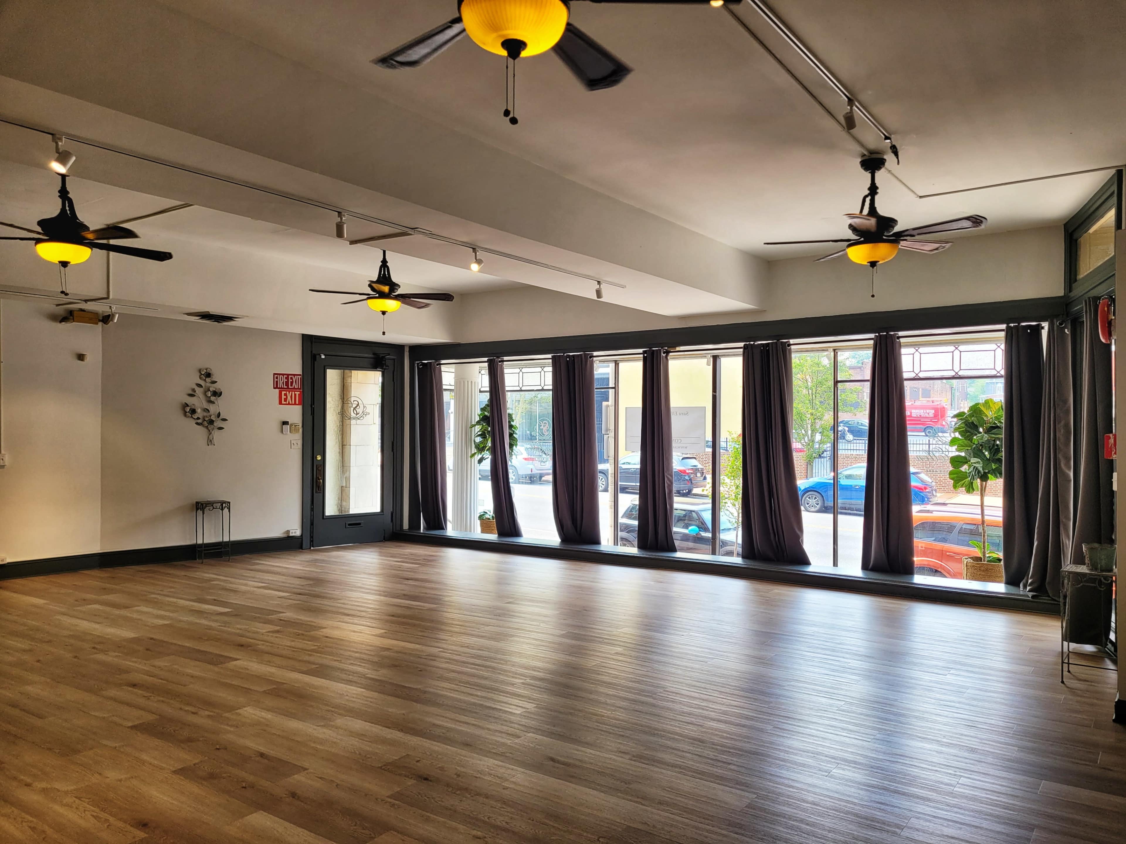 An empty room with wooden flooring, large windows covered by gray curtains, and ceiling fans.