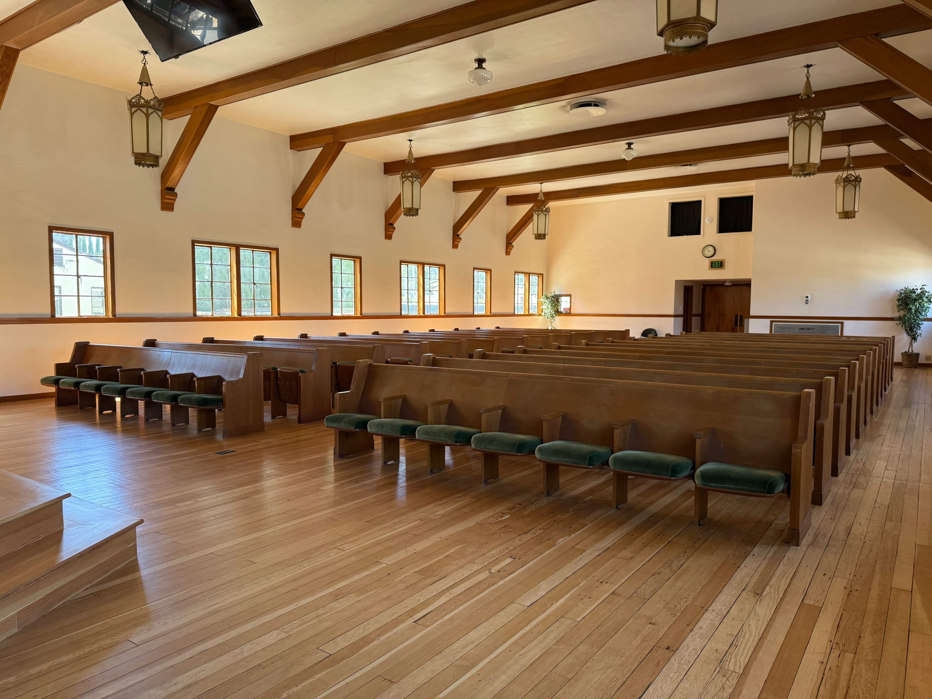 The image shows an empty church interior with wooden pews arranged in rows, wooden beams on the ceiling, and large windows allowing natural light to enter.