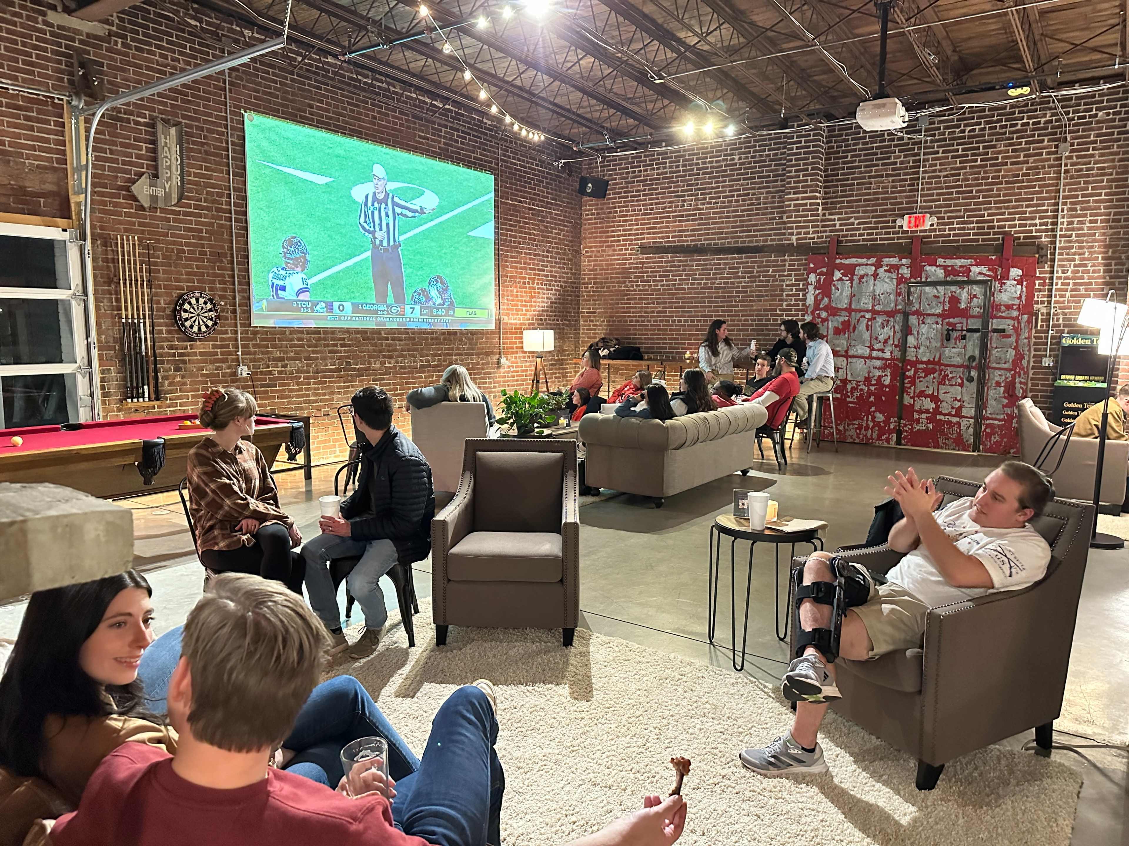 A group of people socializes in a spacious, illuminated room with exposed brick walls while watching a sports game projected on a large screen.