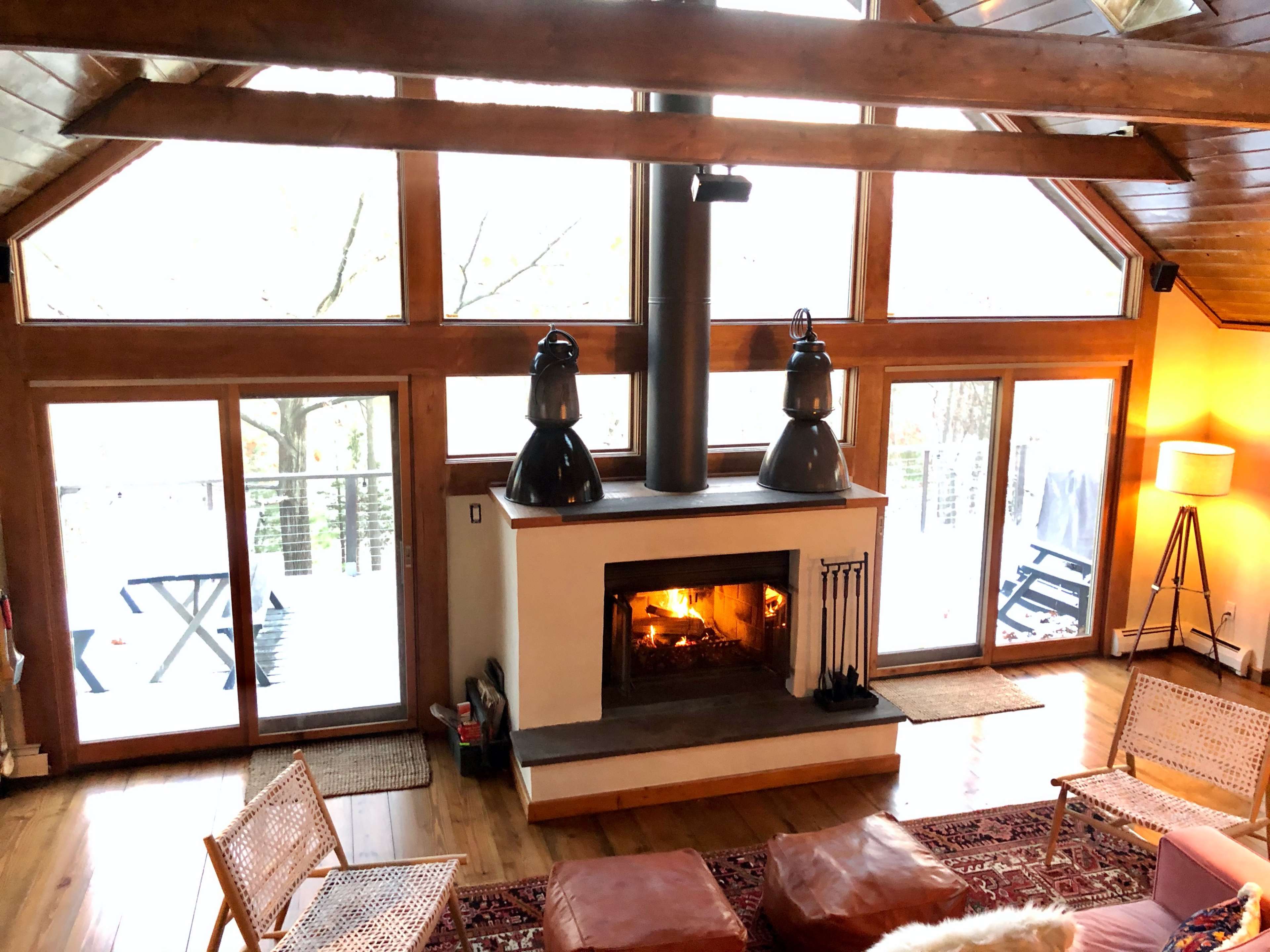A cozy living room with a central fireplace, large windows, wooden beams, and modern furniture.