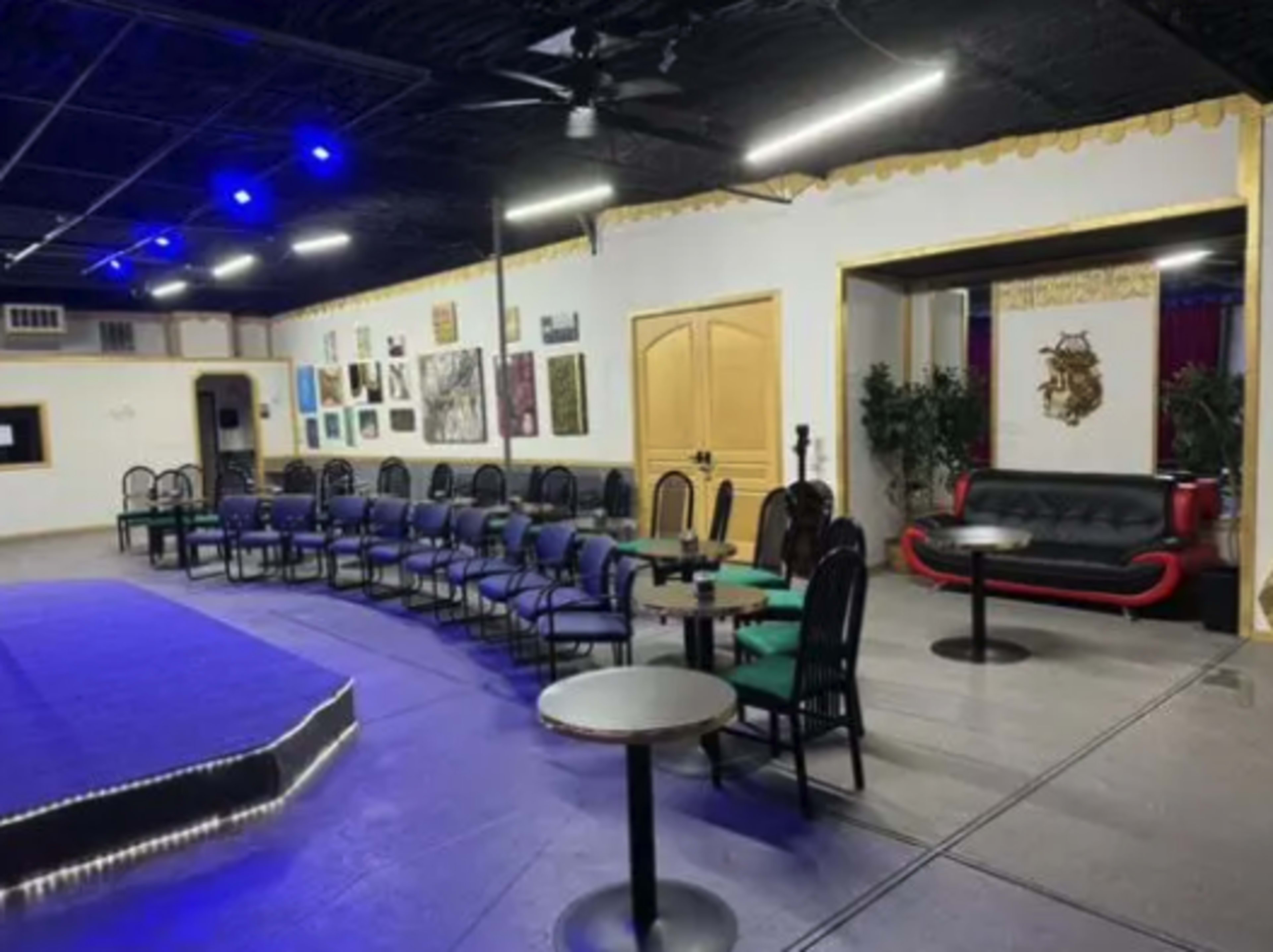An empty performance space with rows of chairs facing a small stage, accompanied by tables and artworks on the walls.