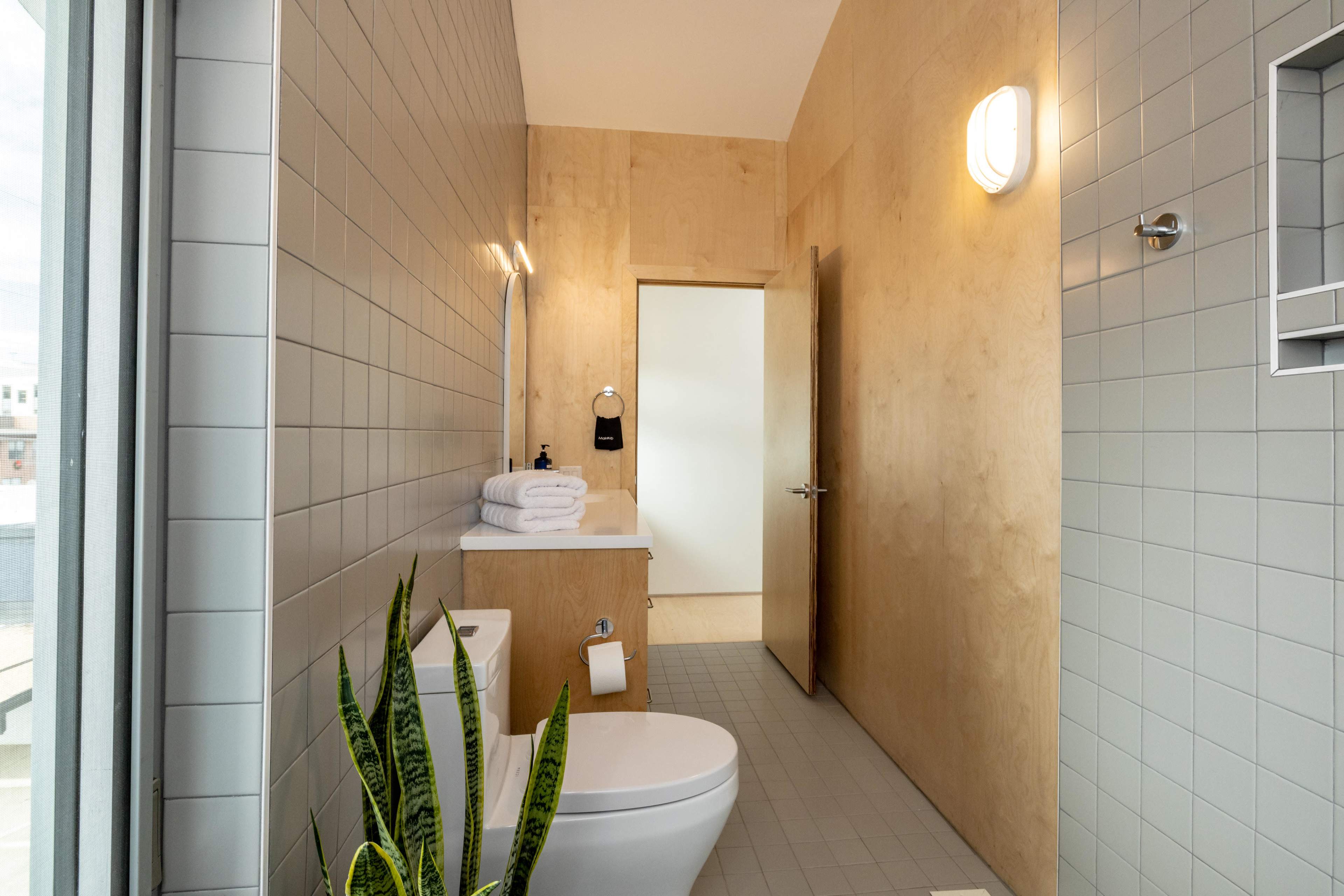 A modern bathroom featuring a toilet, sink with rolled towels, a mirror, and a large window with indoor plants.