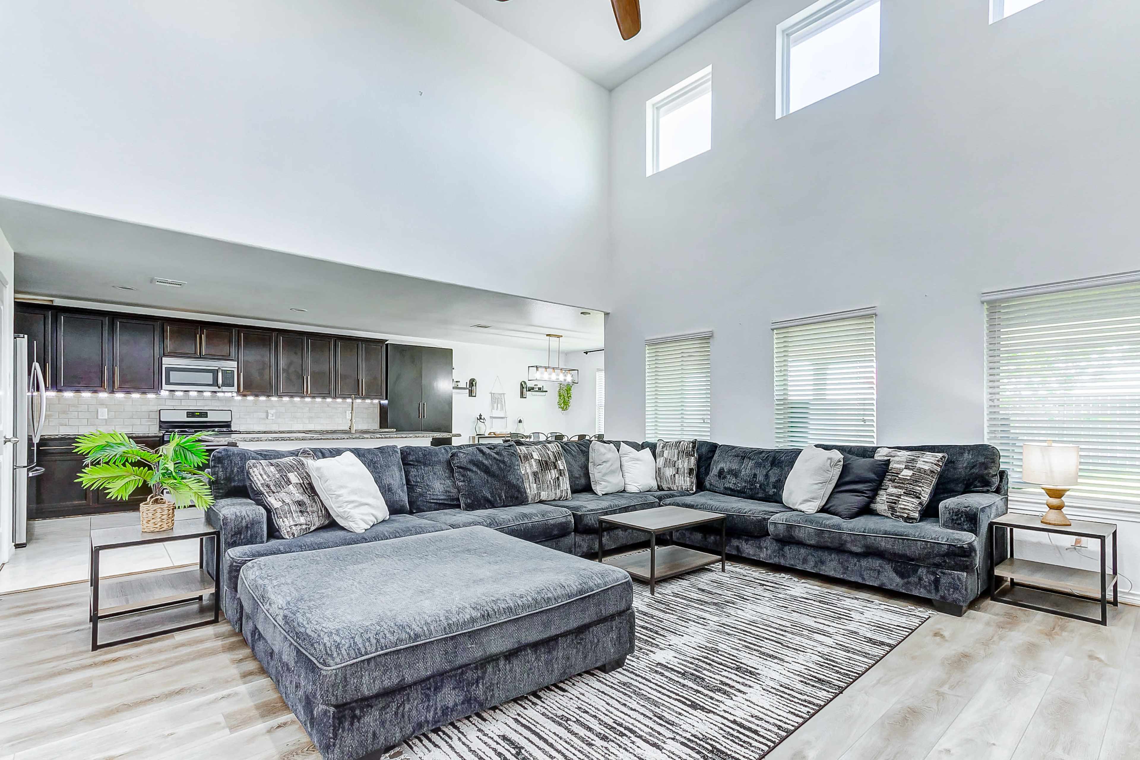A spacious living room features a large sectional sofa, a coffee table, and large windows allowing natural light, adjacent to a kitchen with dark cabinetry.