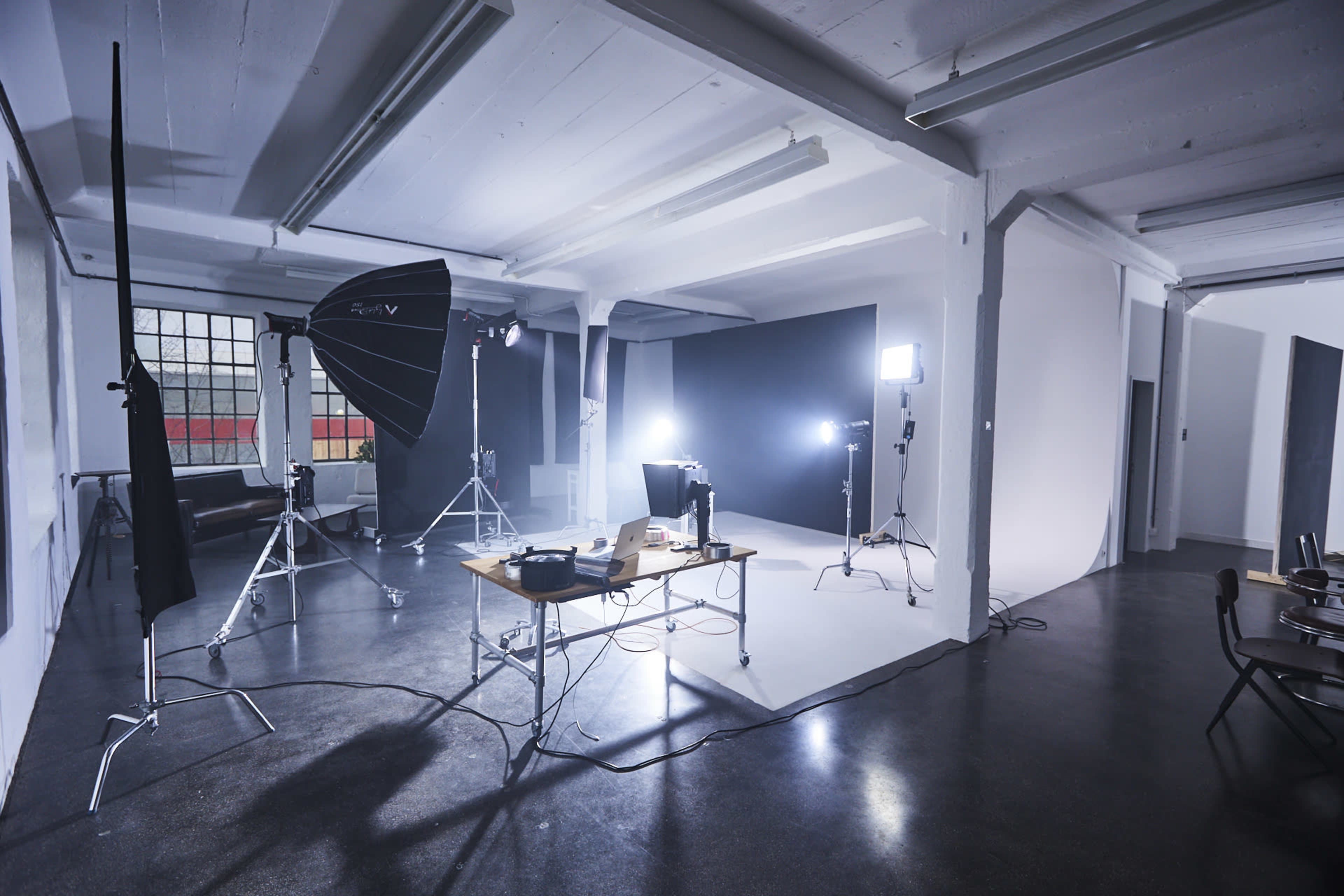 The image shows a photography studio with a table, computer, and lighting equipment set up against a black backdrop.