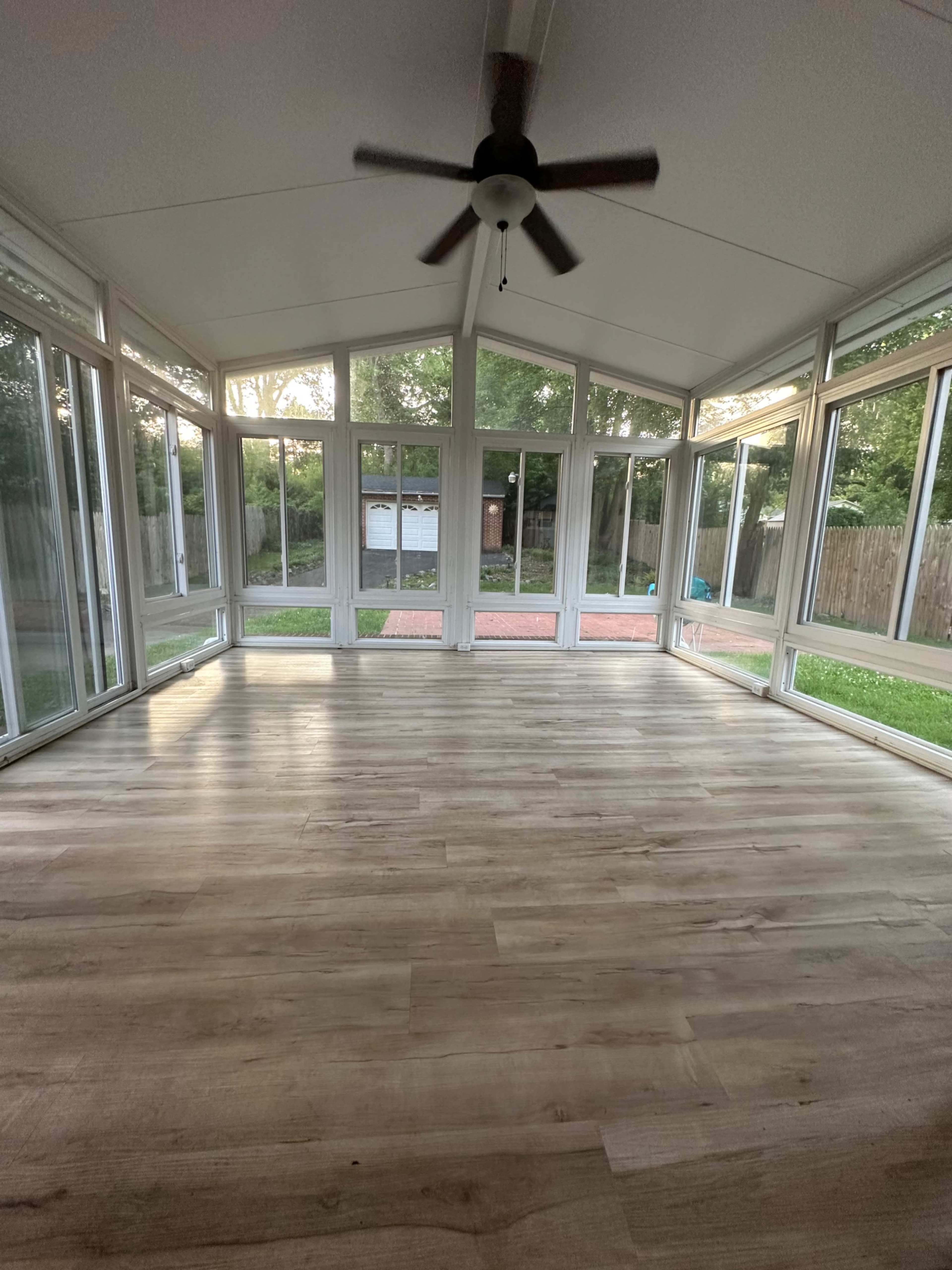 A spacious sunroom with large windows, a ceiling fan, and laminate flooring, overlooking a grassy area and a partially visible outdoor space.