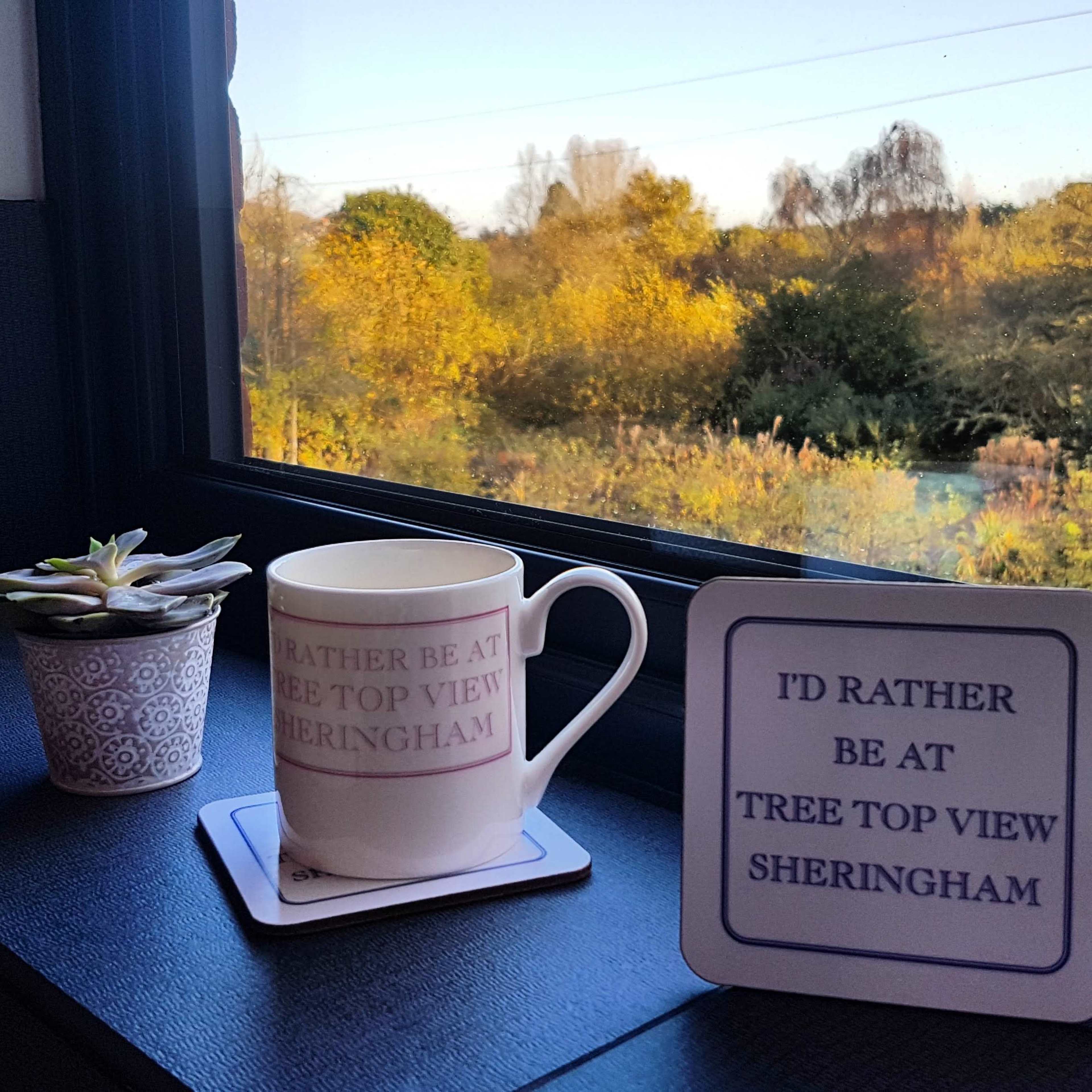 A white mug and a decorative sign are placed on a windowsill next to a succulent, overlooking a landscape with trees and foliage in the background.