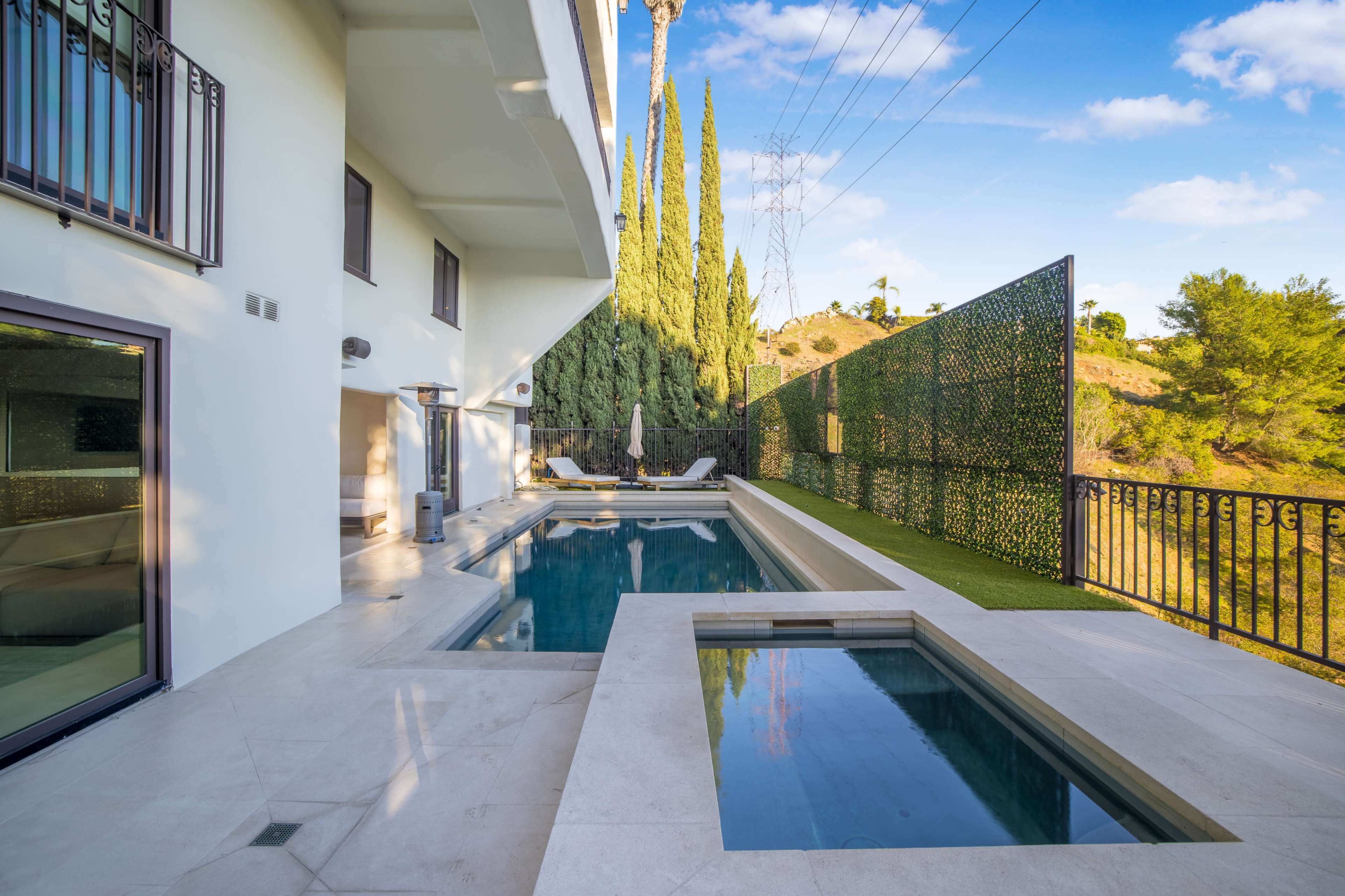 The image shows a modern outdoor area featuring a swimming pool, a hot tub, and landscaped greenery against a hillside backdrop.