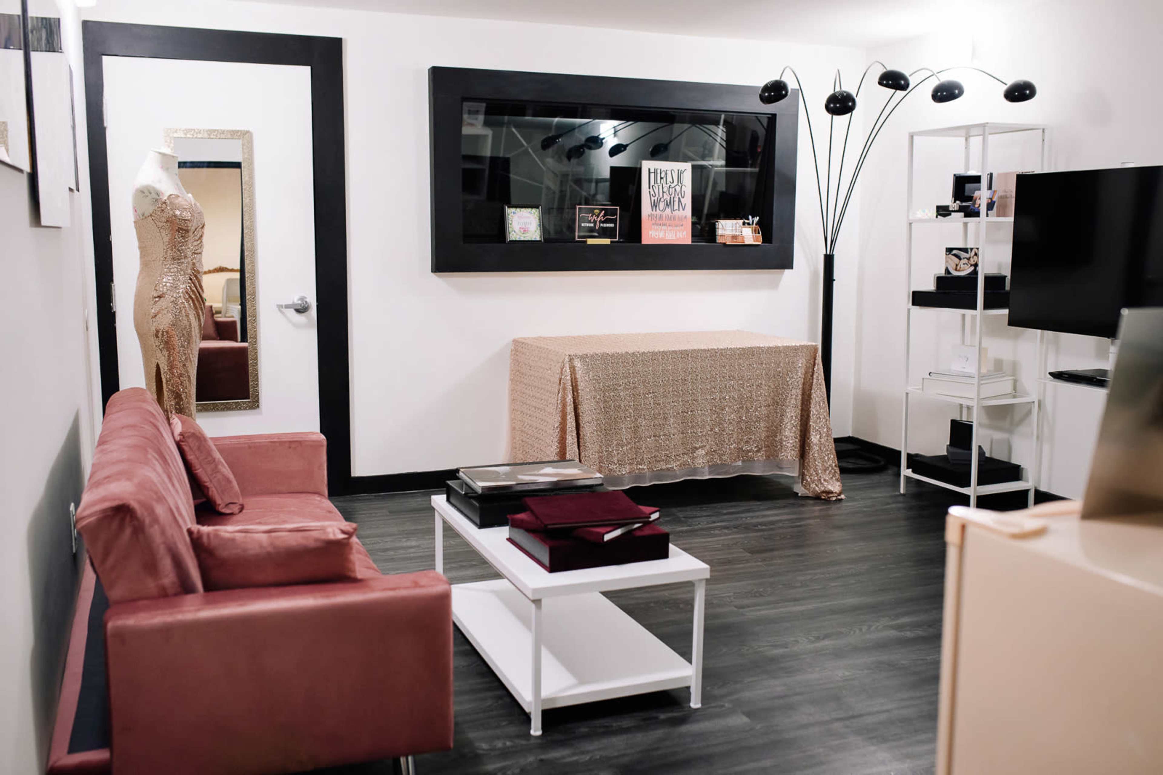 A small, neatly arranged room features a pink sofa, a decorative table with a gold tablecloth, a floor lamp, and shelves displaying various items.