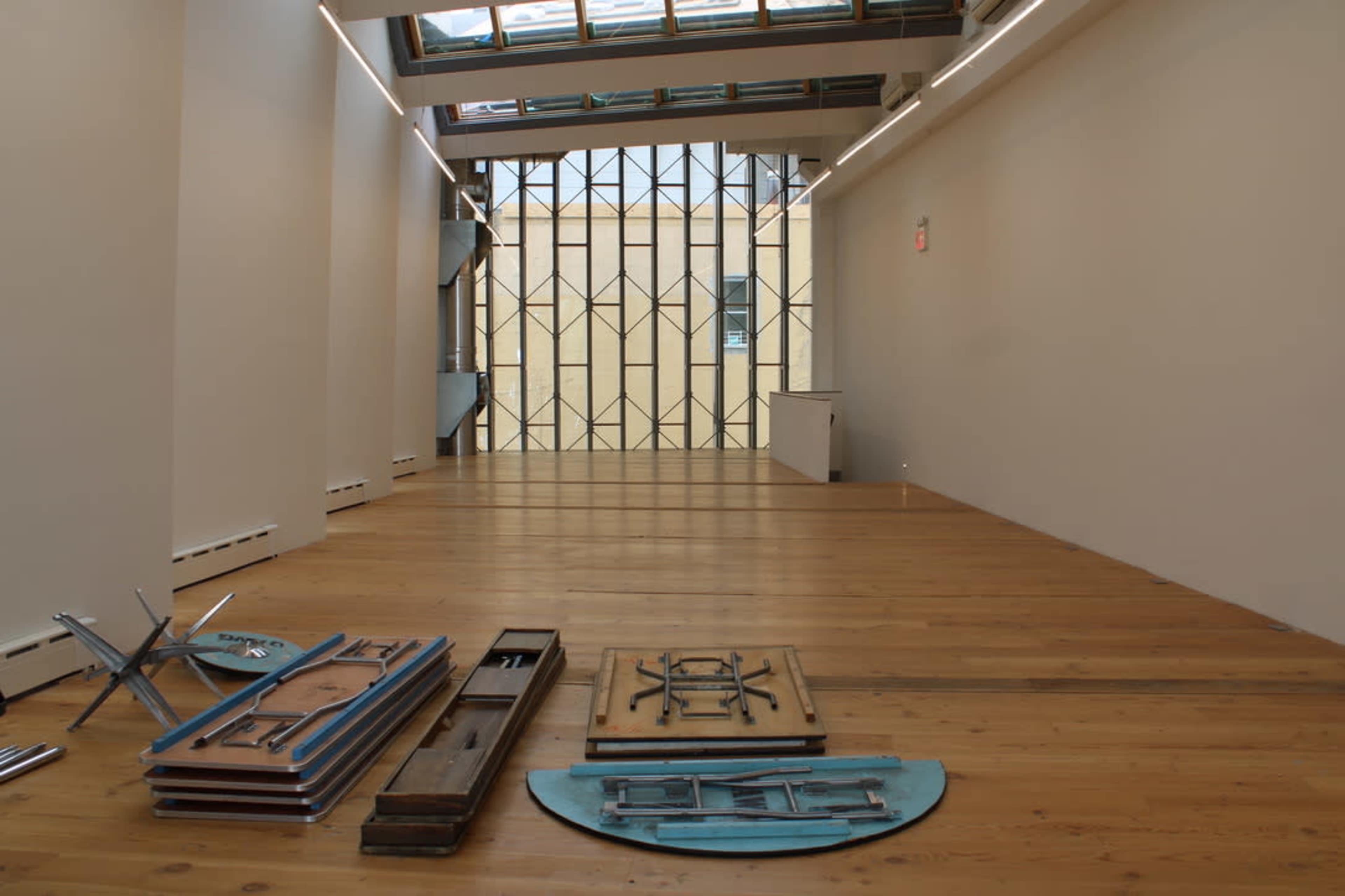The image shows an empty art gallery with wooden floors and a large window structure that allows natural light to enter, alongside some disassembled furniture arranged on the floor.