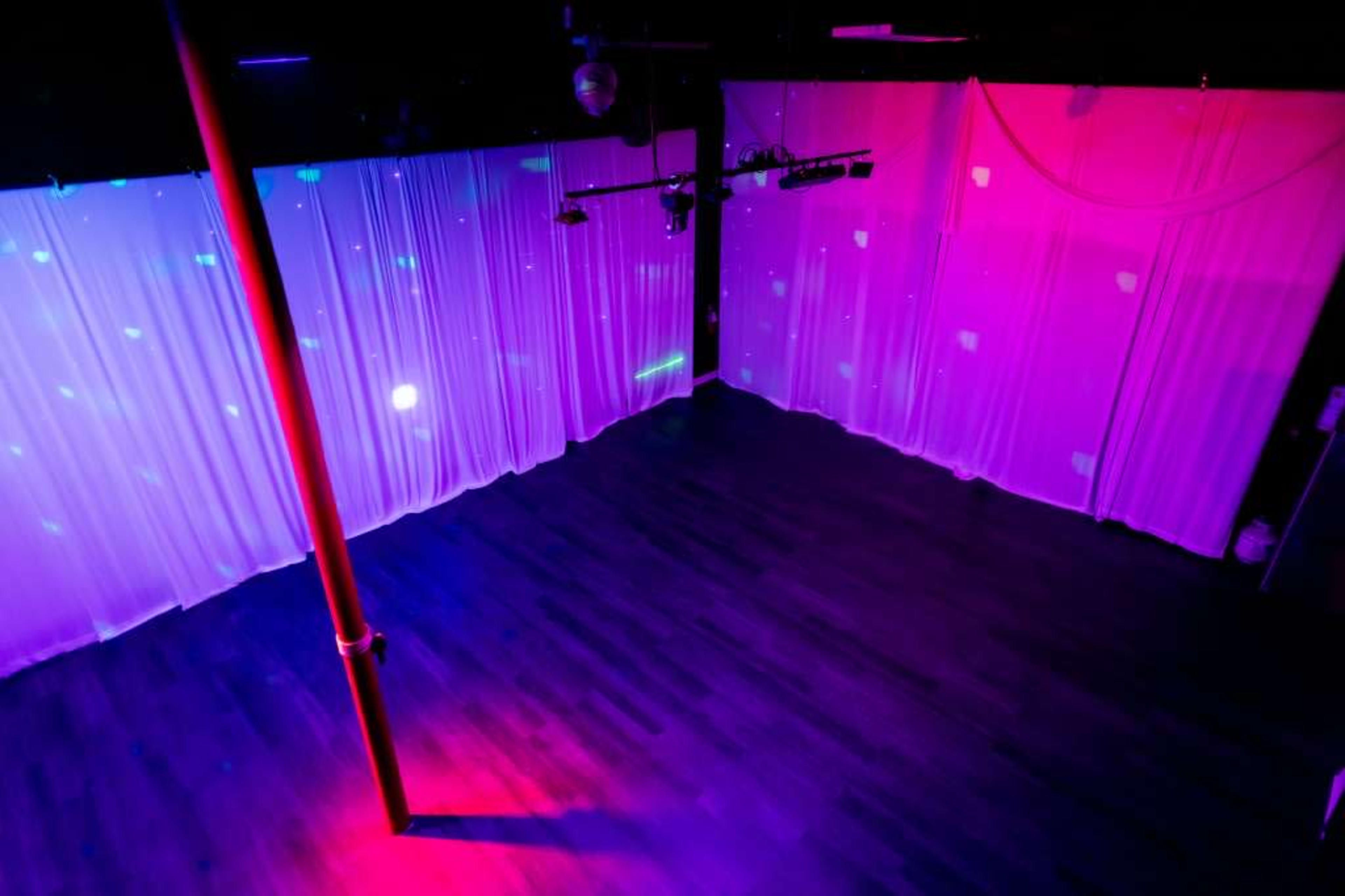 A spacious dance studio features sheer curtains and a polished wooden floor, illuminated by colorful lights.