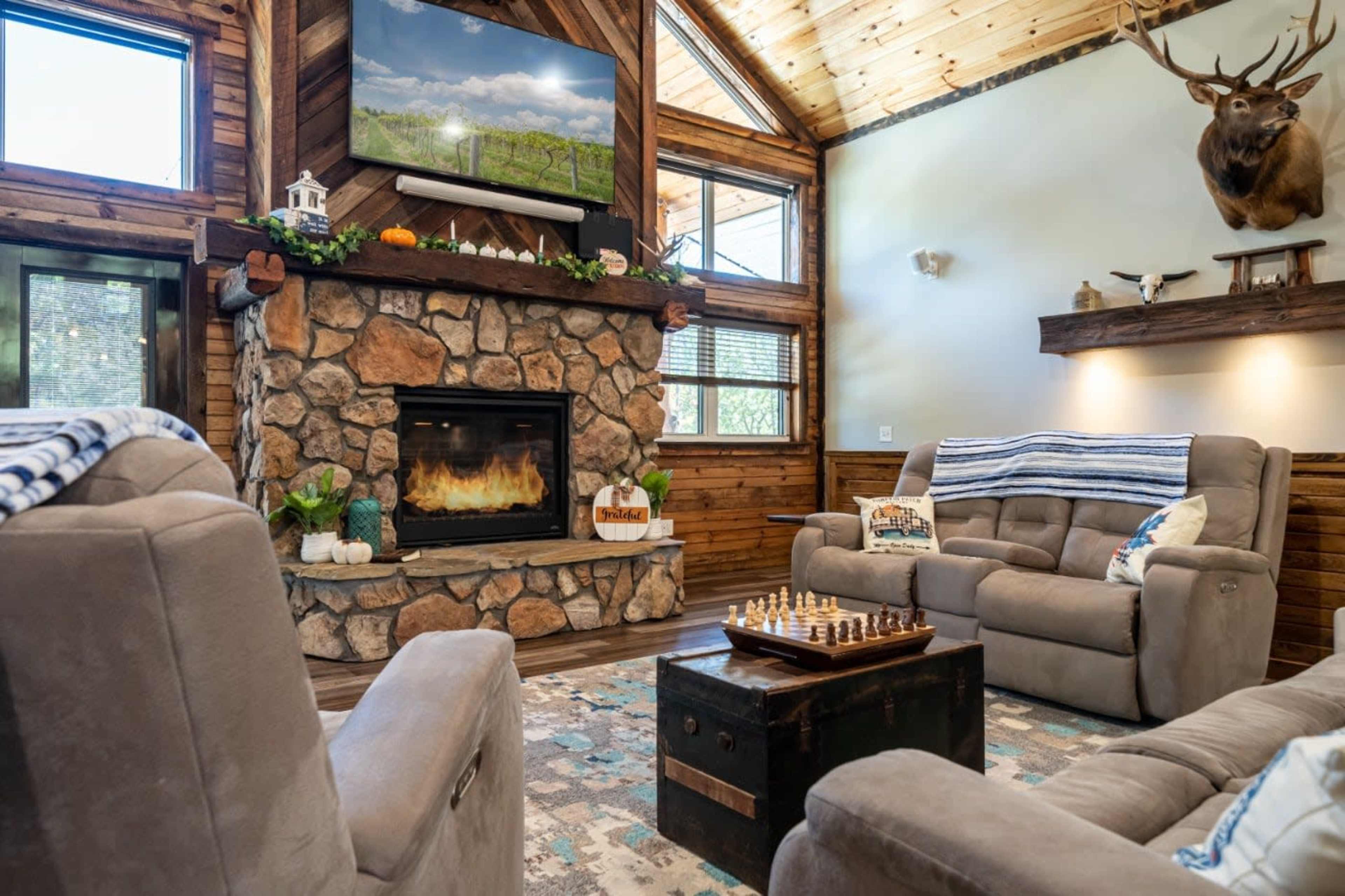 The image depicts a cozy living room featuring a stone fireplace, a mounted deer head, and a chess set on a small table, with wooden walls and a television mounted above the fireplace.