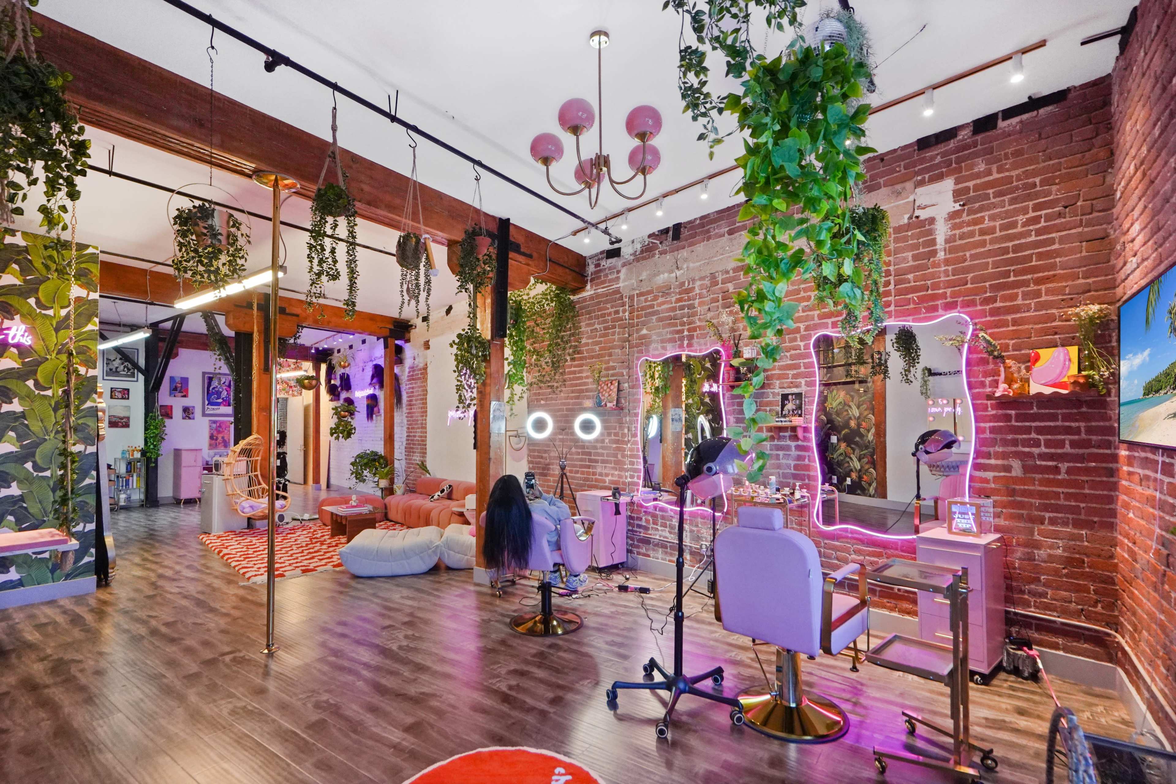 A brightly lit studio space features exposed brick walls, hanging plants, and various makeup stations with neon lighting.