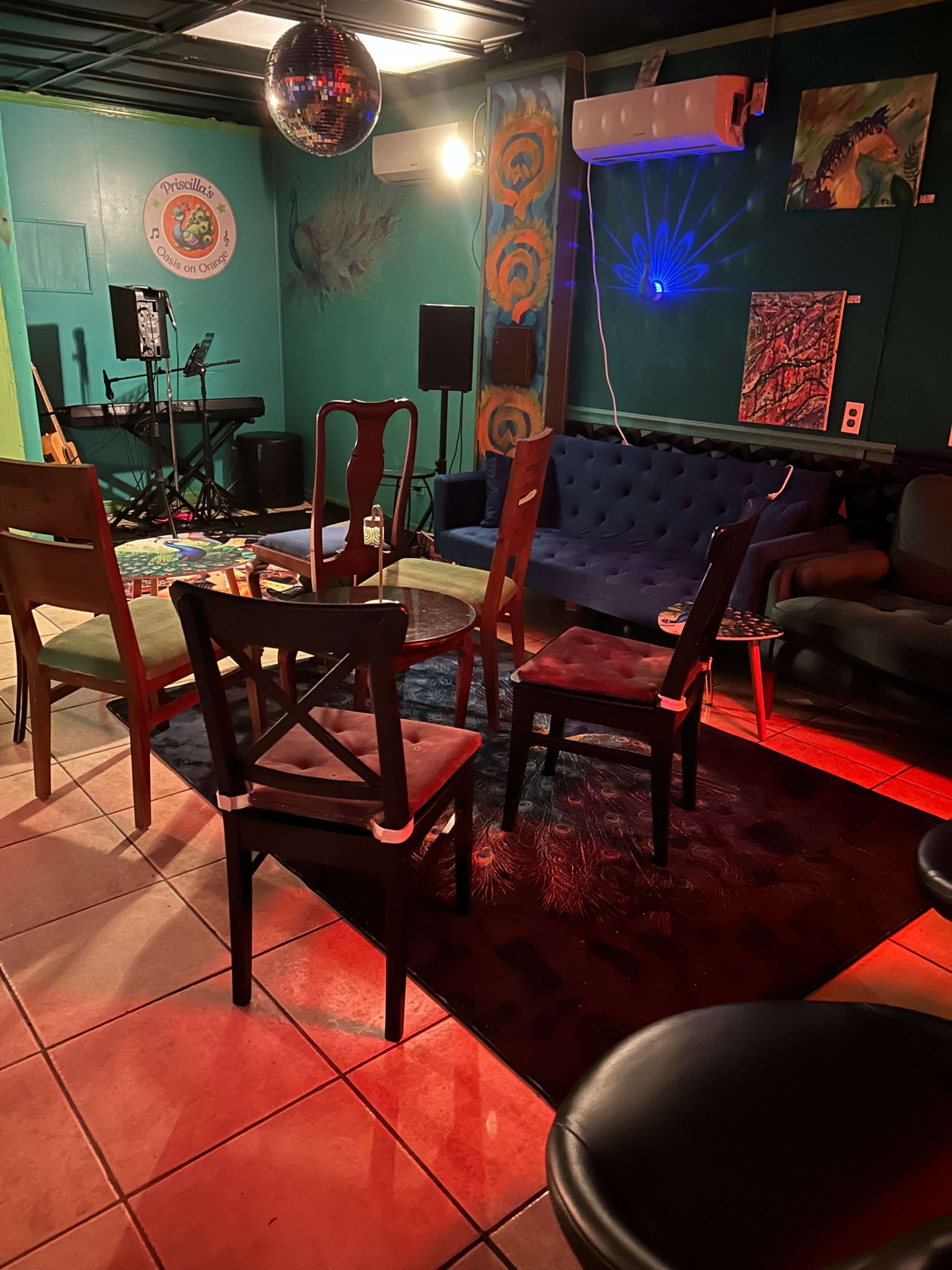 The image shows a cozy lounge area with a blue couch, several chairs, and a stage setup featuring musical equipment in a room with colorful wall art and low lighting.