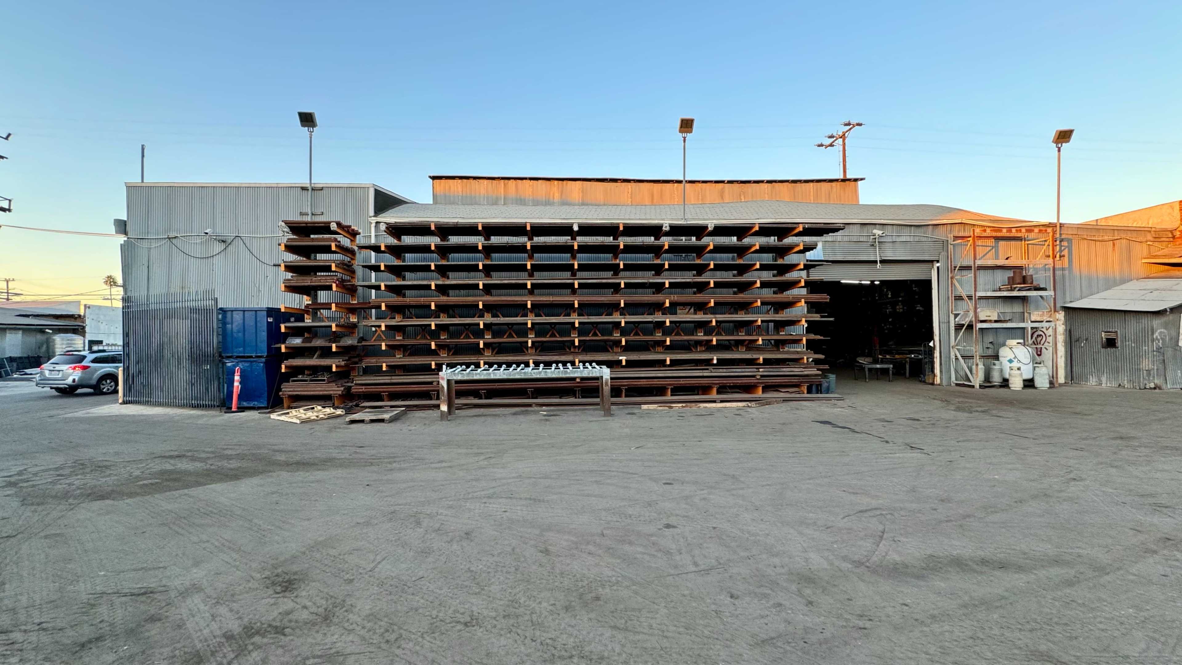 A warehouse exterior features a large stacked structure of wooden beams next to an open garage entrance on a gravel lot.