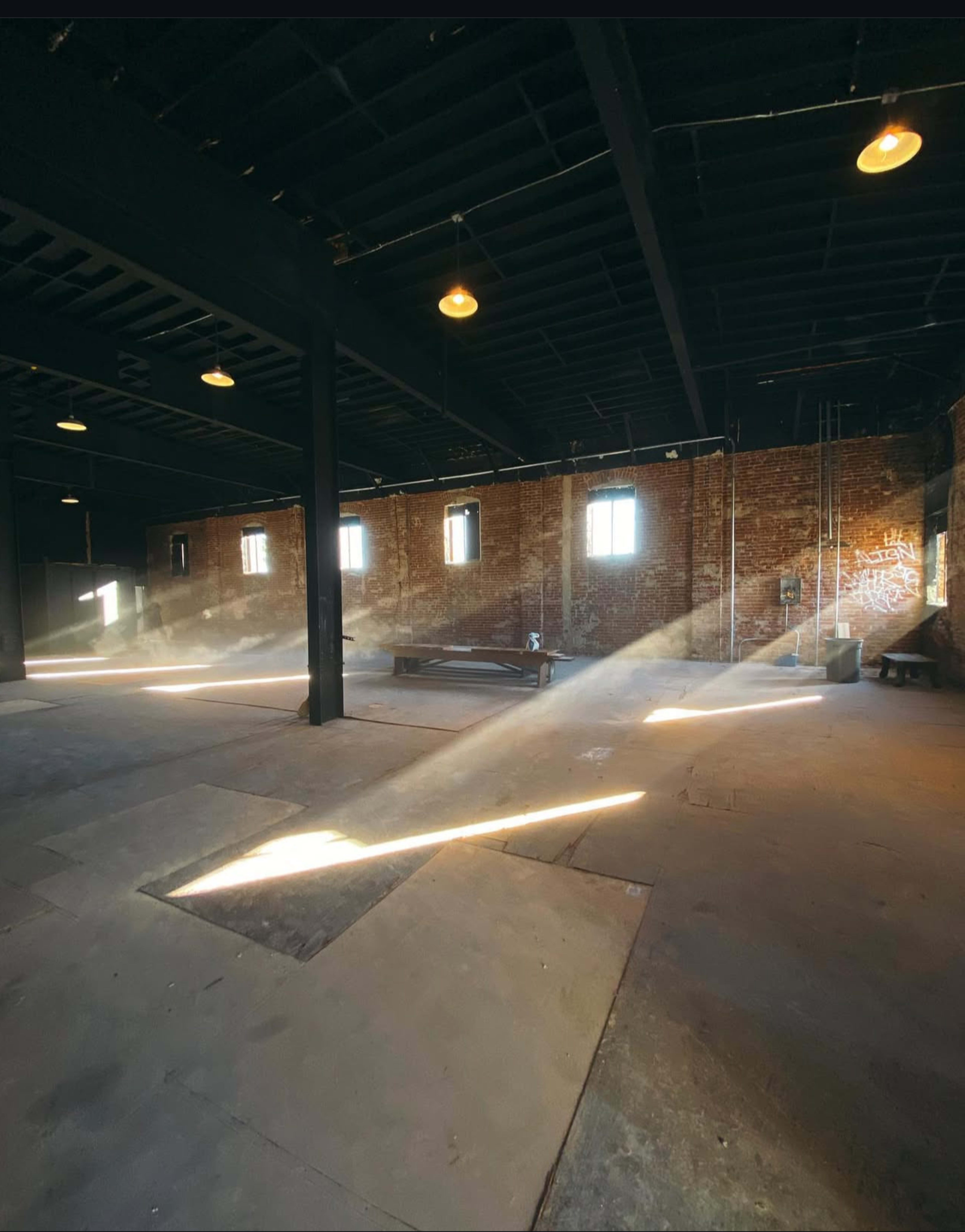 A sunbeam shines through the windows of an empty industrial space with brick walls and a concrete floor.