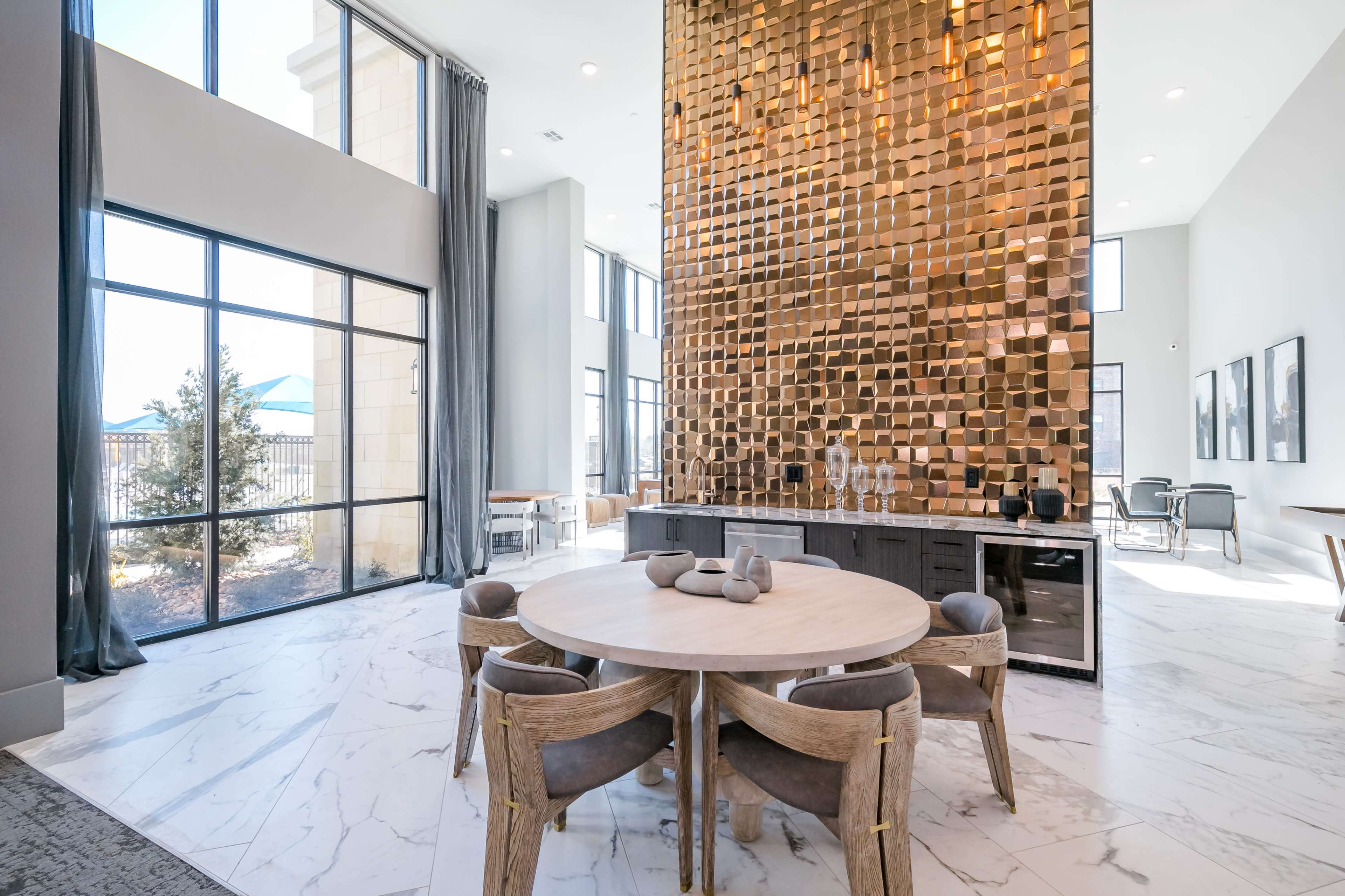 A spacious modern room features a large round wooden table surrounded by upholstered chairs, with a striking gold-accented wall and large windows letting in natural light.