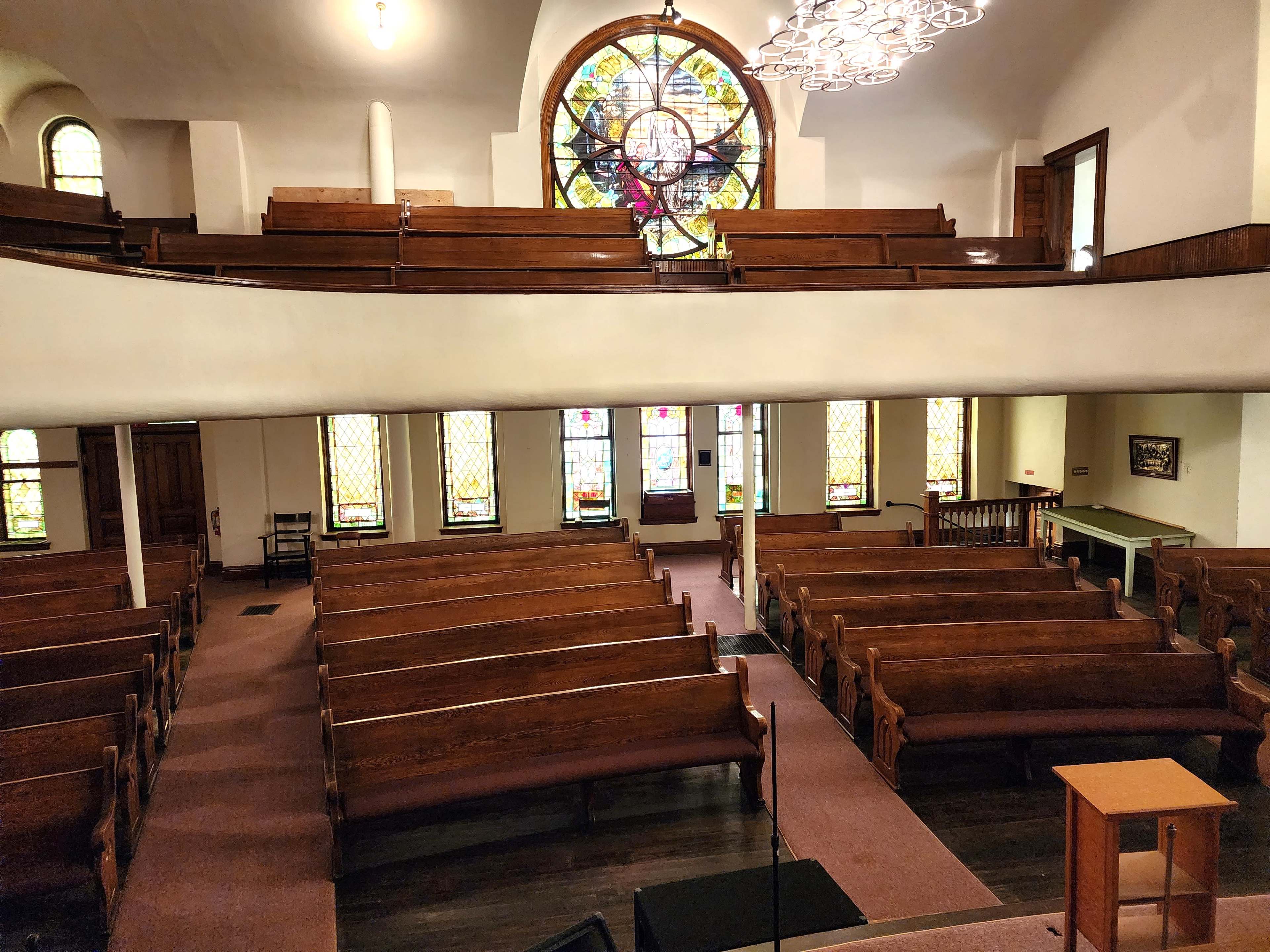 The image shows the interior of a church with wooden pews arranged around a central aisle and stained glass windows illuminating the space.