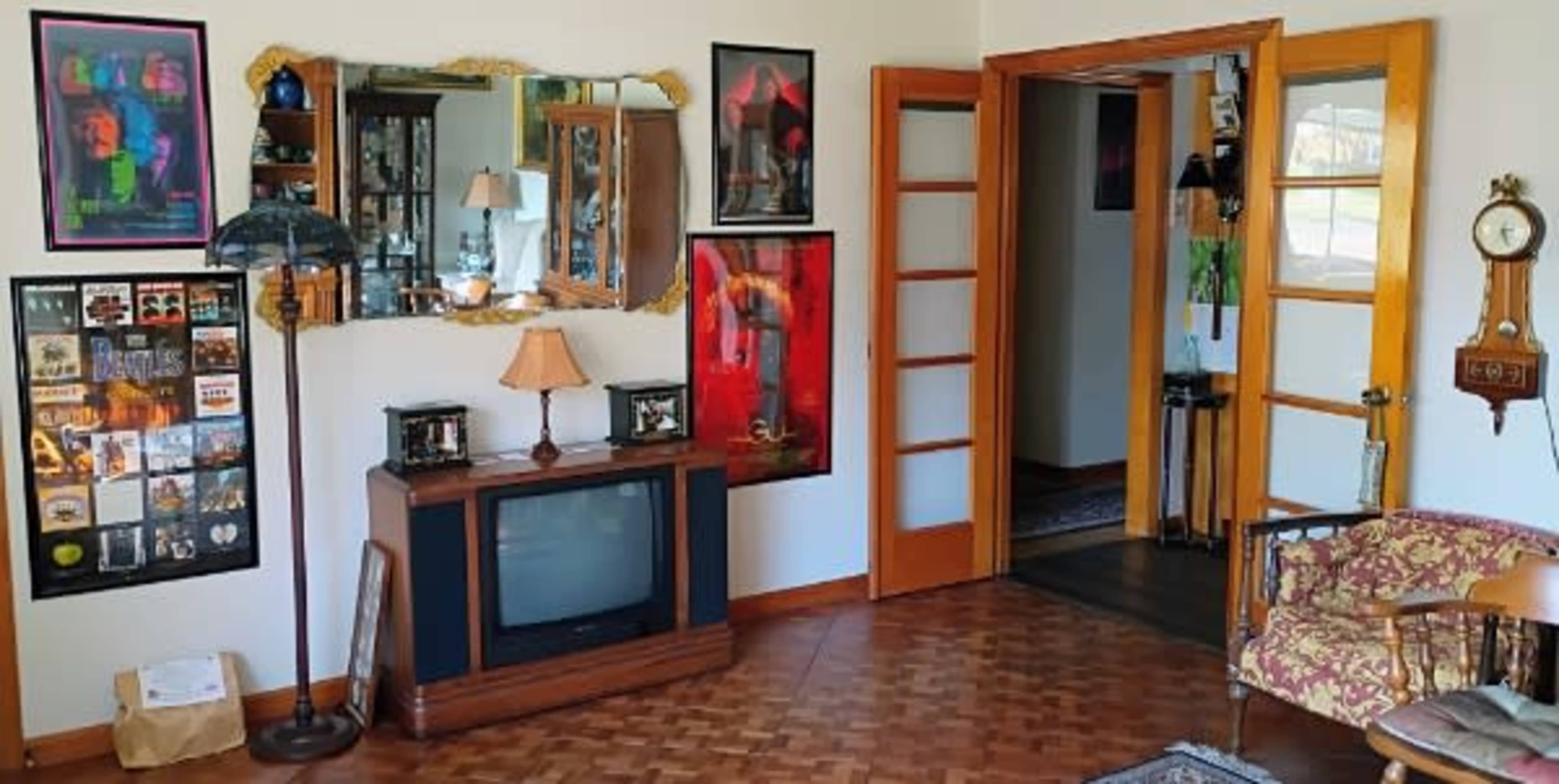 The image shows a living room with a vintage television, a decorative lamp, framed artwork on the walls, and wooden doors leading to other areas.