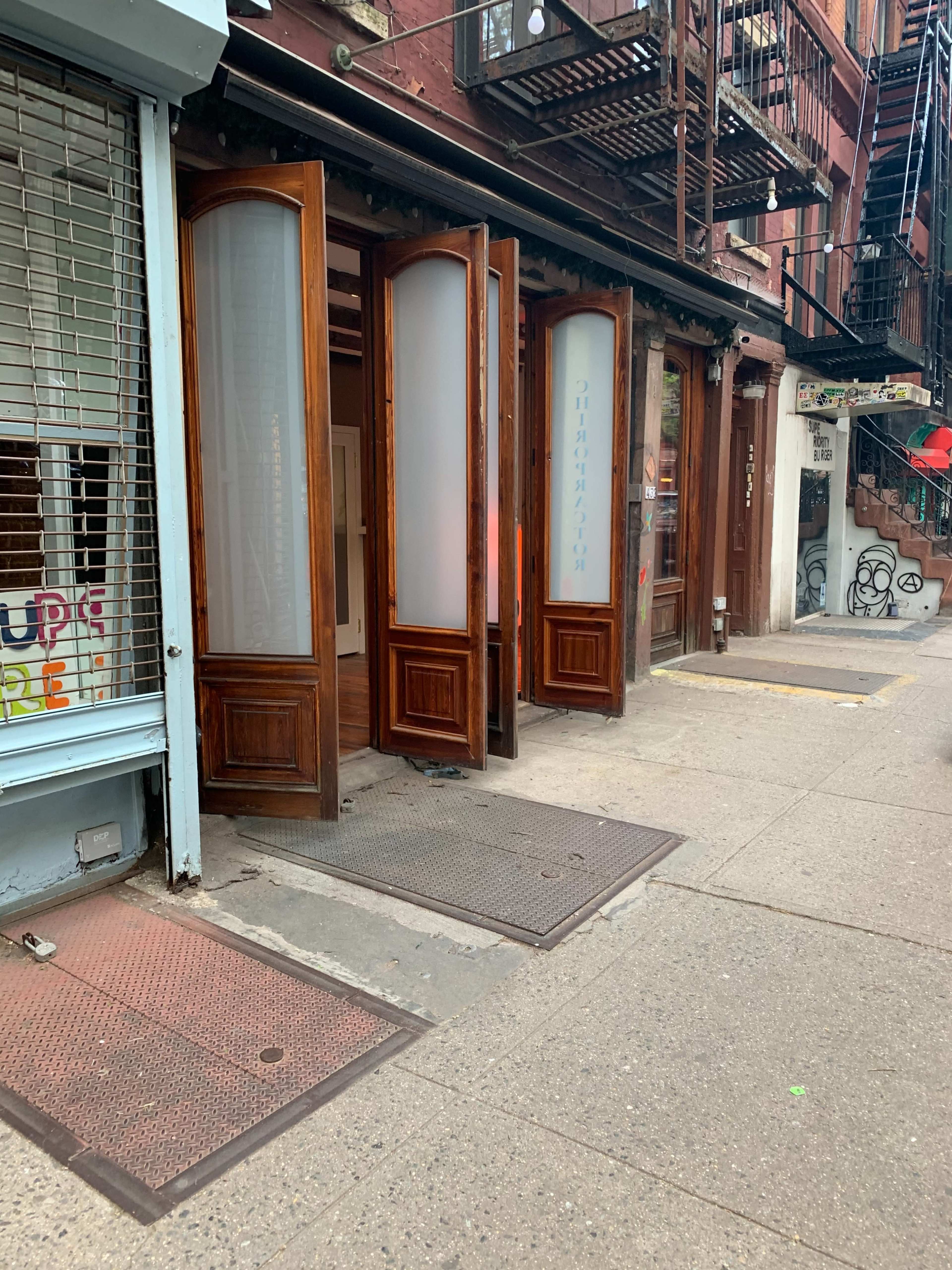 A sidewalk with three open wooden doors leading to a building, next to a shop with a closed metal roller shutter.
