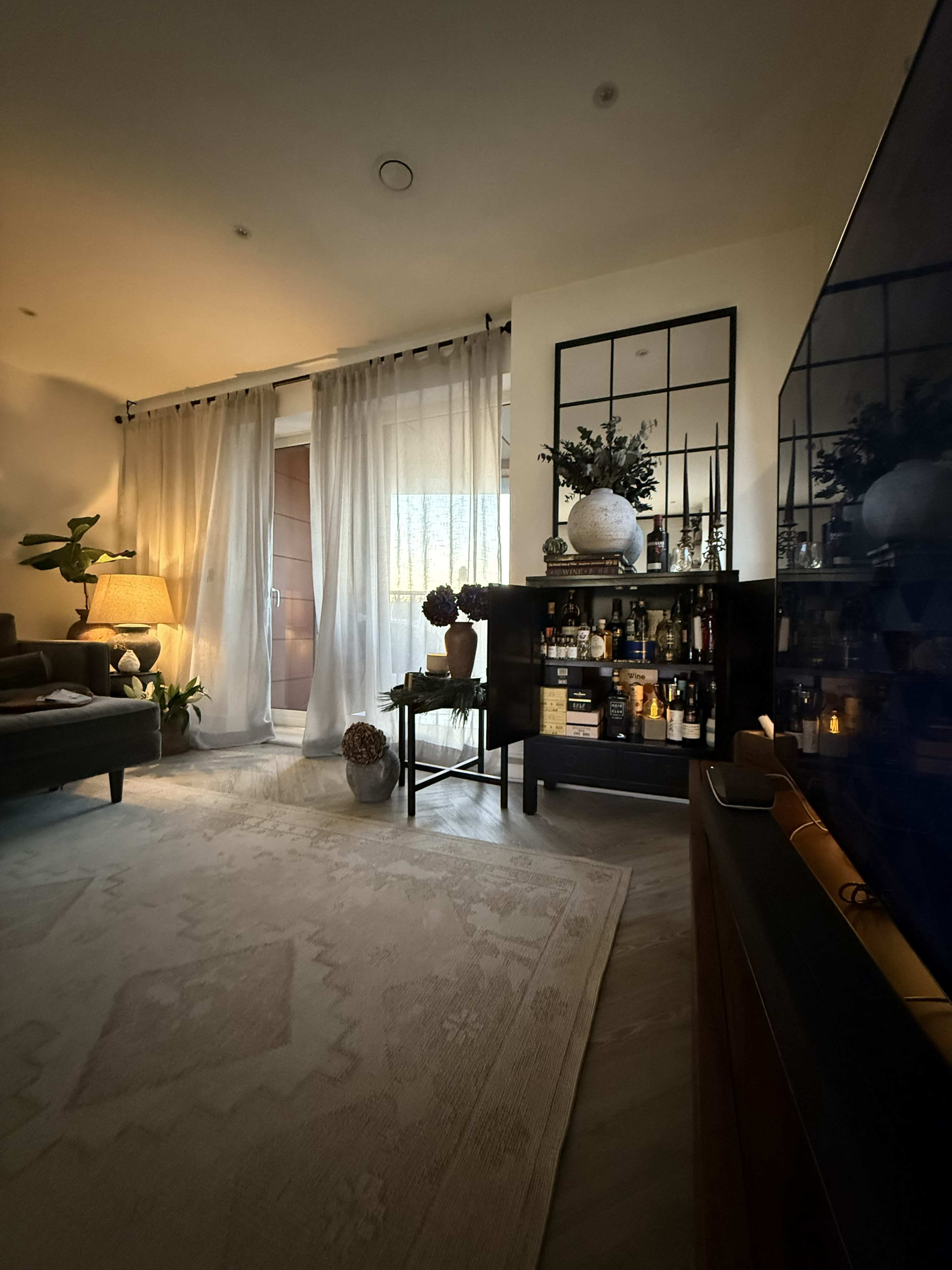 The image features a cozy living room with a seating area, a stylish bar cart, and sheer curtains covering a glass door.