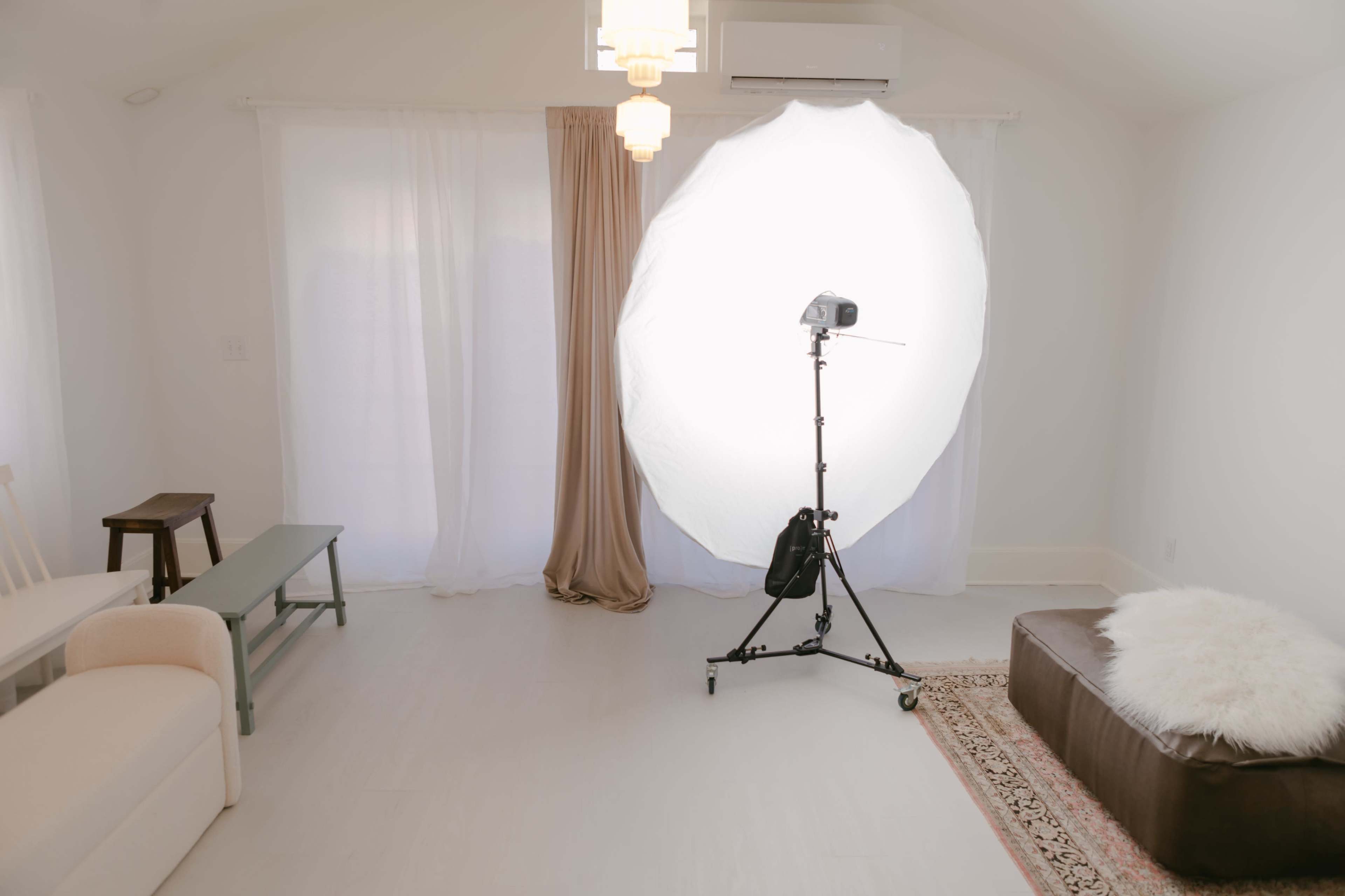 A bright, minimalistic interior space featuring a softbox lighting setup, a cream-colored sofa, a green table, and a brown leather ottoman with a white fur throw.