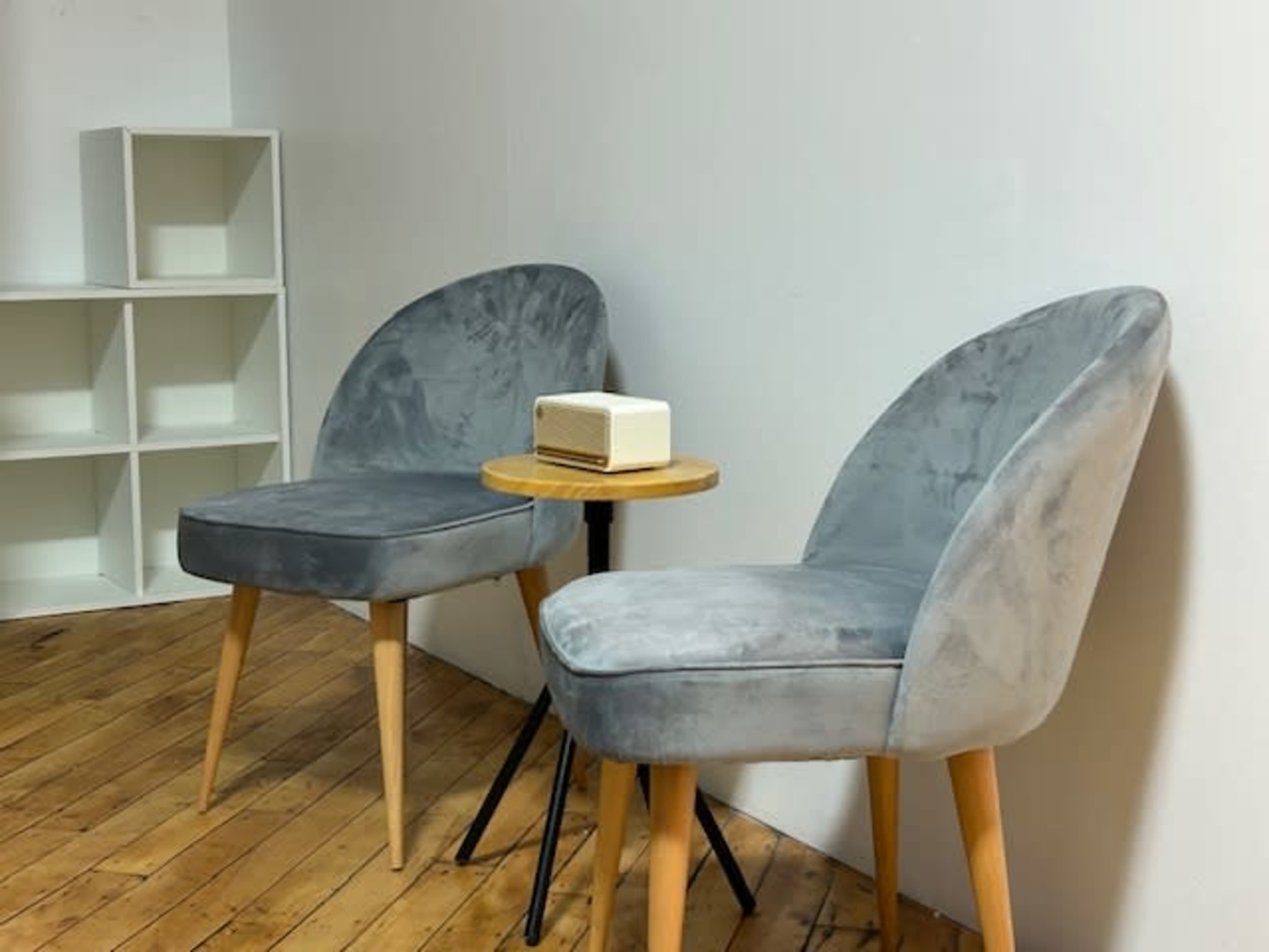 The image shows two gray upholstered chairs with wooden legs positioned near a small round table and a white shelf against a plain wall.
