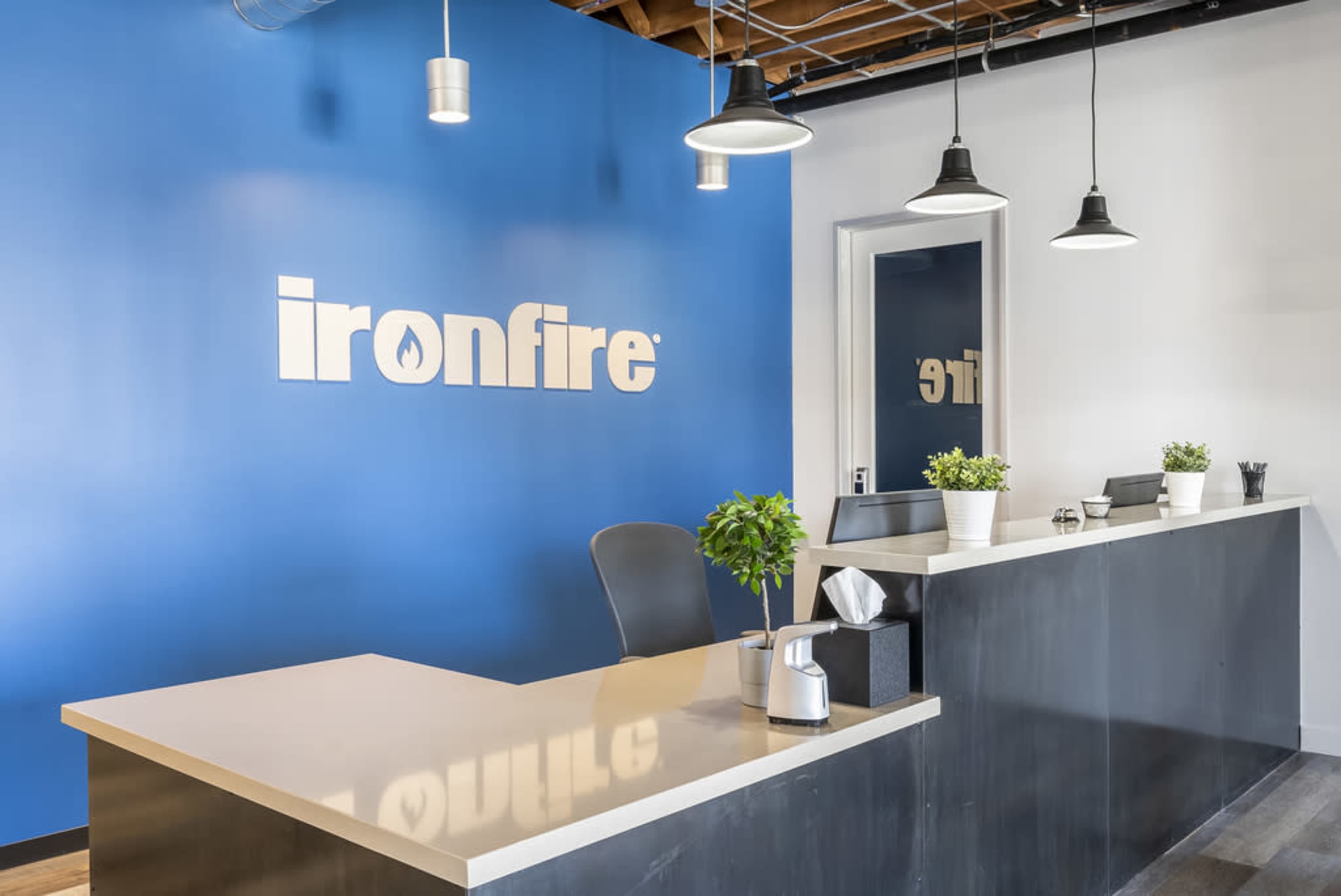 The image shows a modern reception area with a blue wall displaying the "ironfire" logo, a sleek counter, and hanging light fixtures.